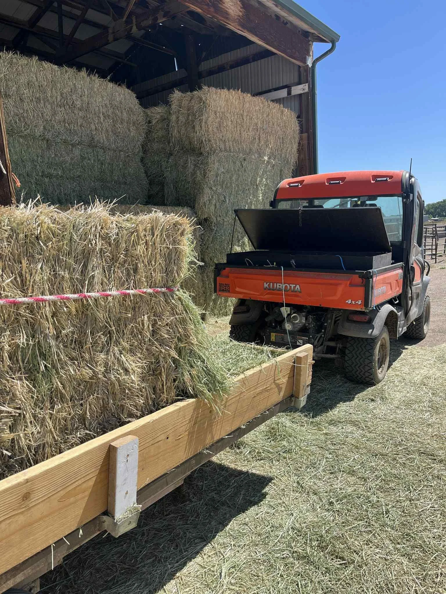 A small utility vehicle with an orange and gray body parked next to a wooden trailer filled with hay bales. The trailer is attached to a barn or shed with a metal roof. The hay bales are stacked inside a shed with a partially open door, and the scene