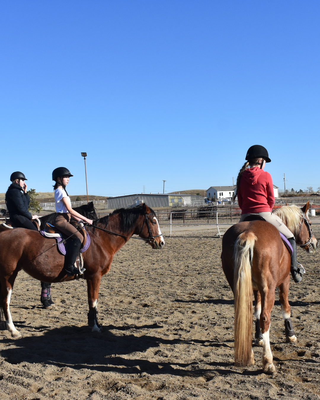Three young women riding horses in an outdoor riding arena on a sunny day.