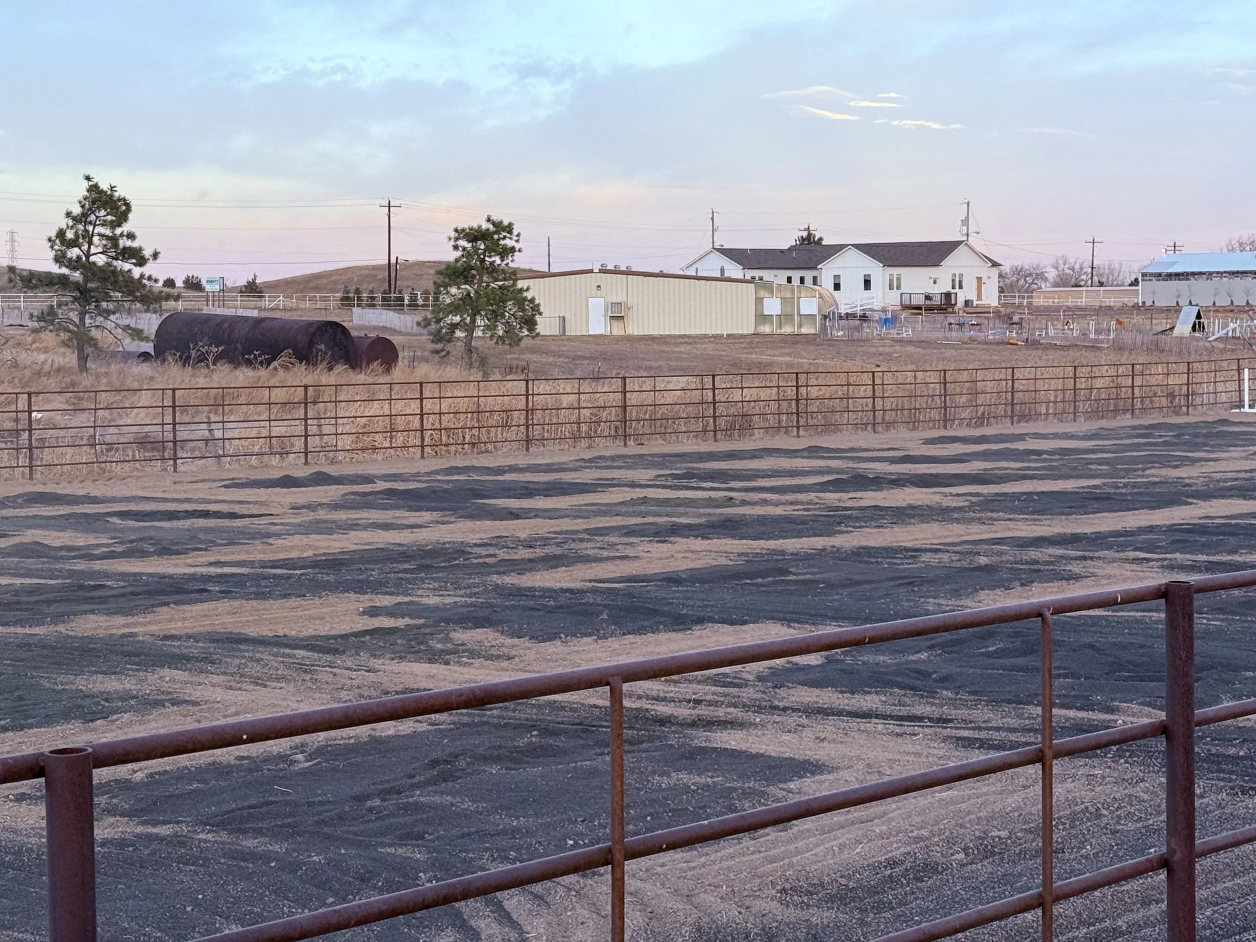Rodeo arena with a dirt surface and a metal fence in the foreground, surrounded by open land, trees, and farm buildings in the background during sunset or sunrise.