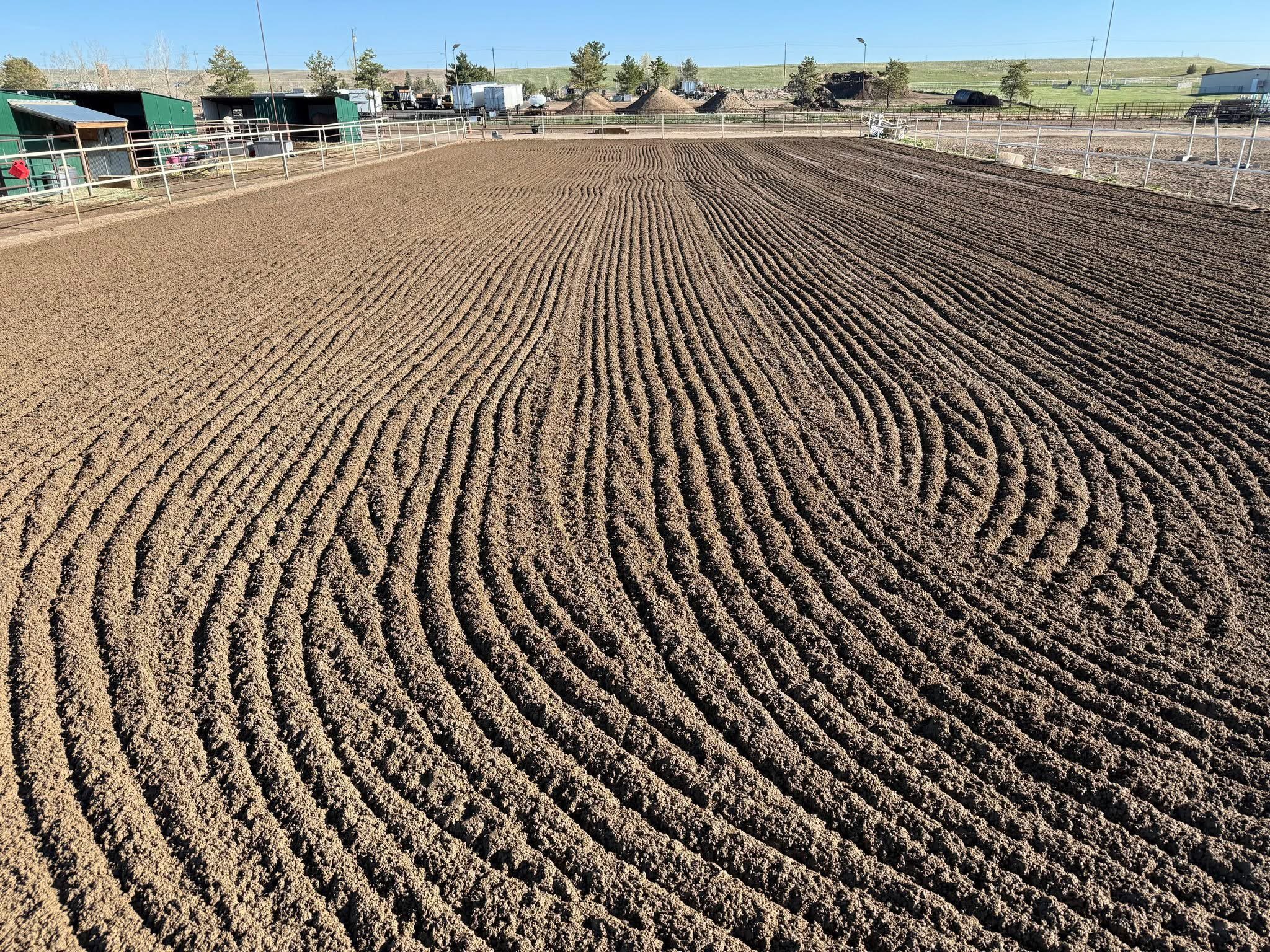 A freshly tilled horse riding arena with mare circles firmed into the dirt, enclosed by white fencing and surrounded by farm buildings and equipment under a clear blue sky.