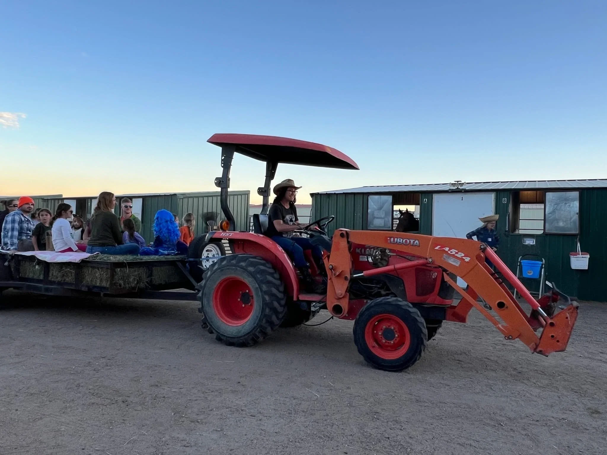 People seated on a flatbed trailer attached to a red tractor, at what appears to be a farm or rural event during dusk, with green barn-like buildings in the background.