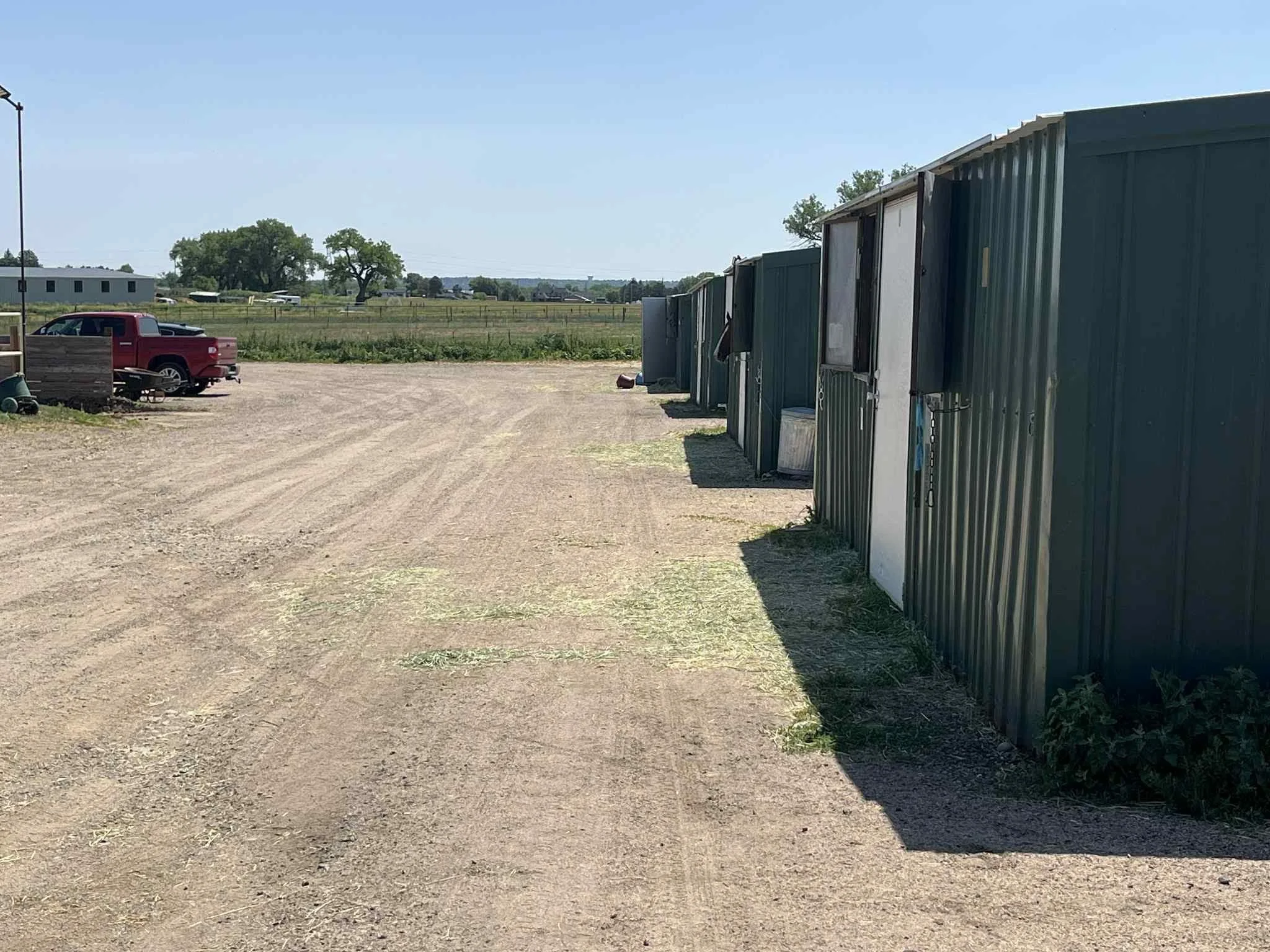 A row of metal storage units or sheds on a dirt lot with a pickup truck parked towards the left and a vast open field with trees in the background under a clear sky.