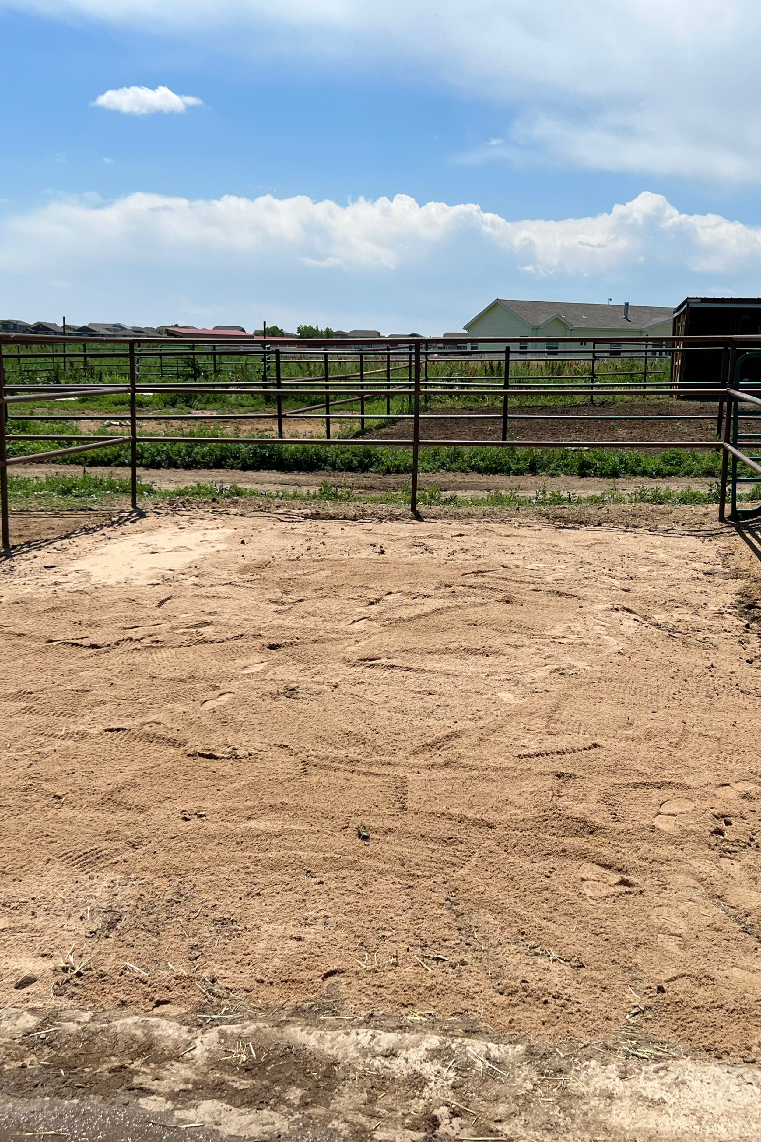 An empty dirt riding arena with metal fencing, green fields, and houses in the background under a partly cloudy sky.