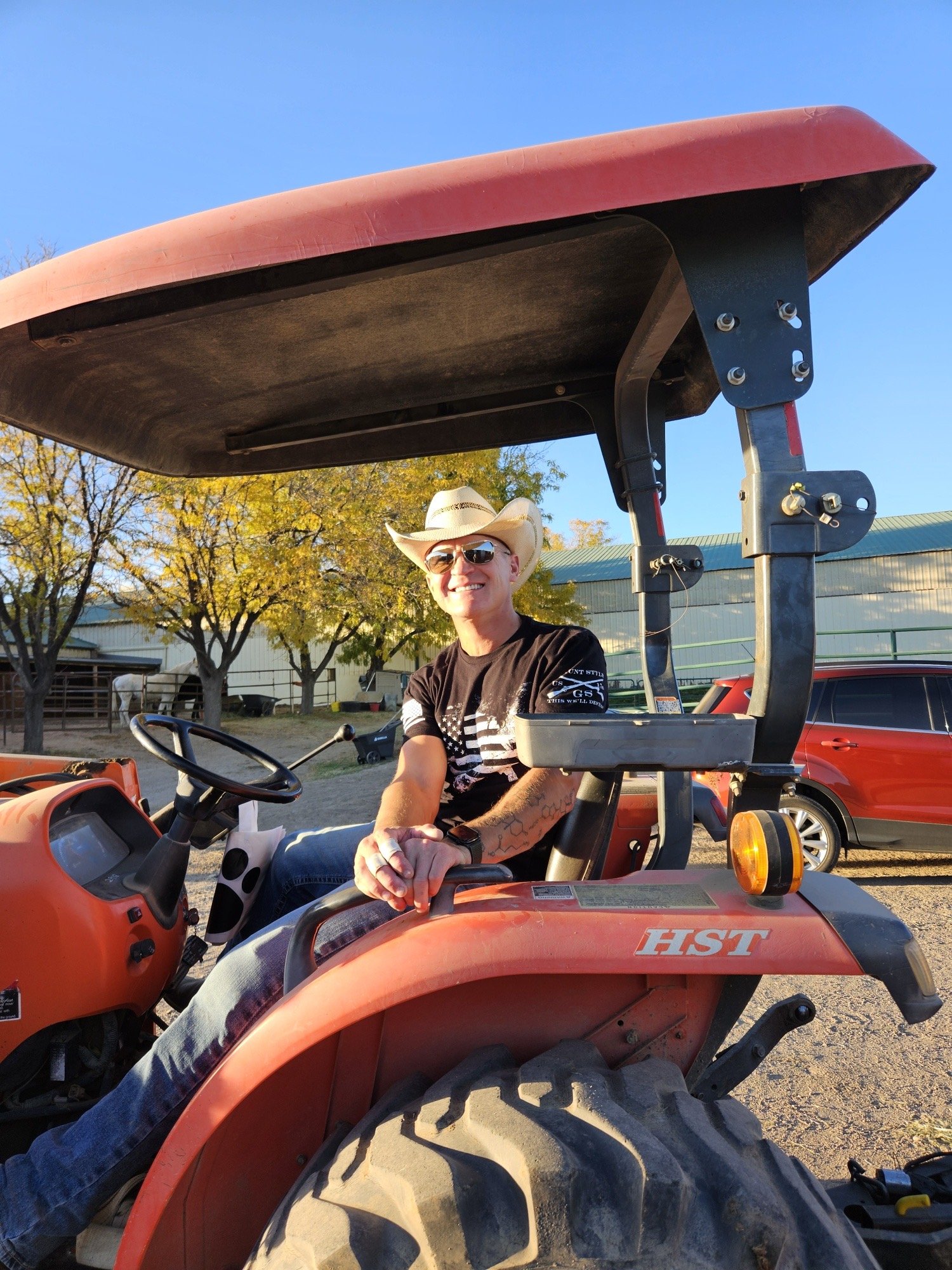 A man wearing a cowboy hat, sunglasses, and a black t-shirt sitting on a red tractor outdoors with trees and a building in the background.