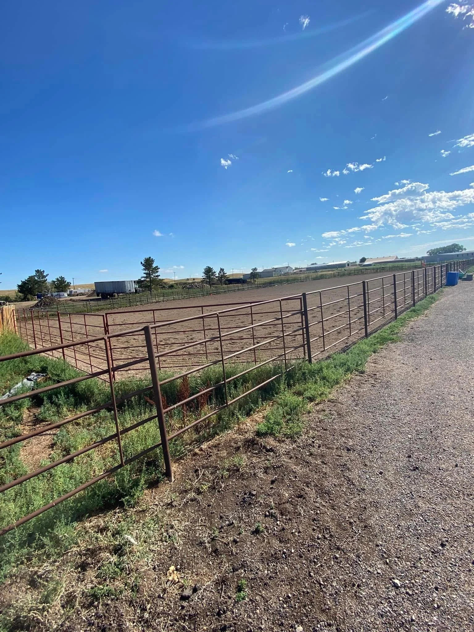 An outdoor scene with a vast, empty dirt arena, surrounded by a metal fence, under a bright blue sky with scattered clouds and lens flare.