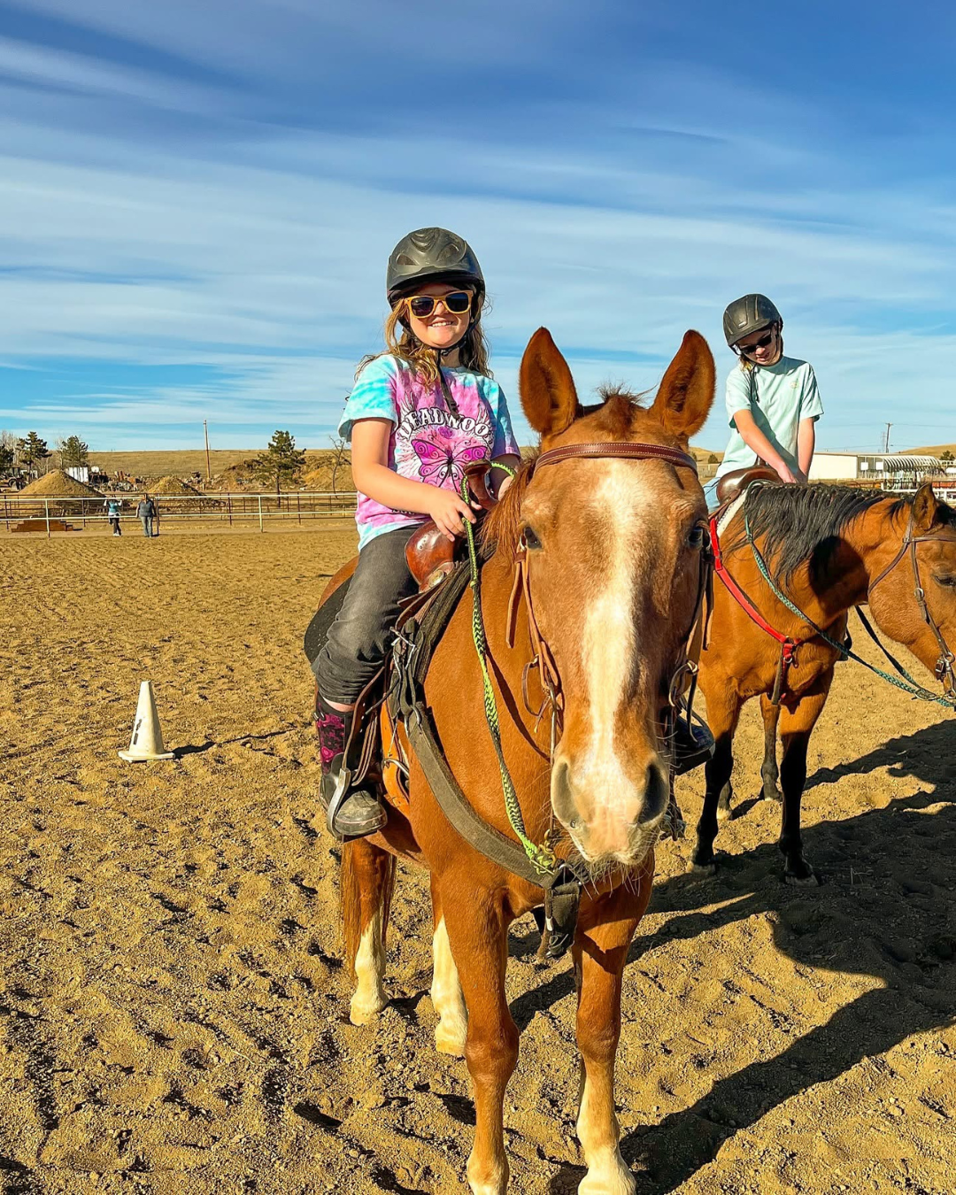 Two children riding horses on a dirt riding arena, with a clear blue sky overhead and a few onlookers in the background.