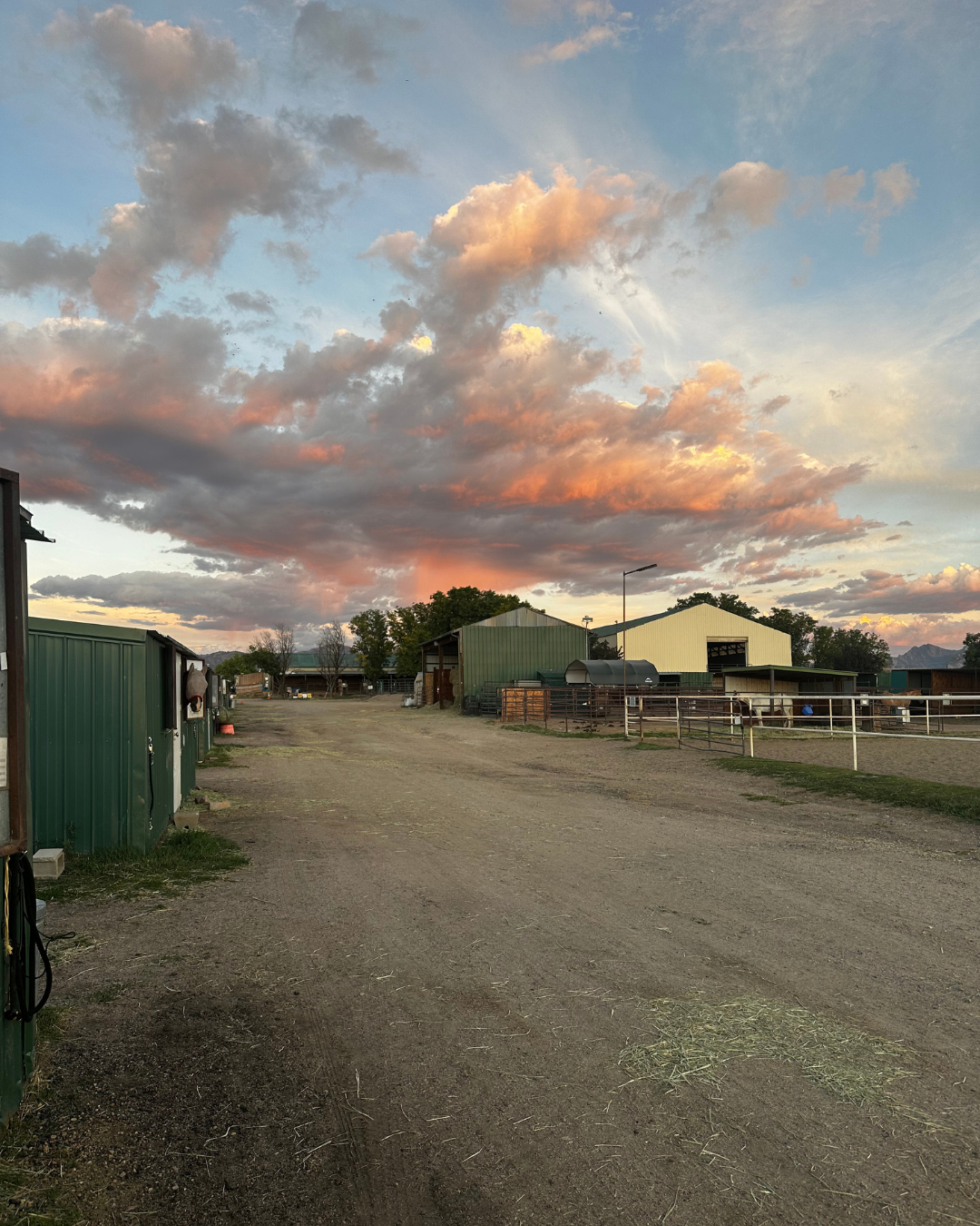 A rural farm scene during sunset with colorful clouds in the sky, dirt road, green and white barns, and fencing for animals.