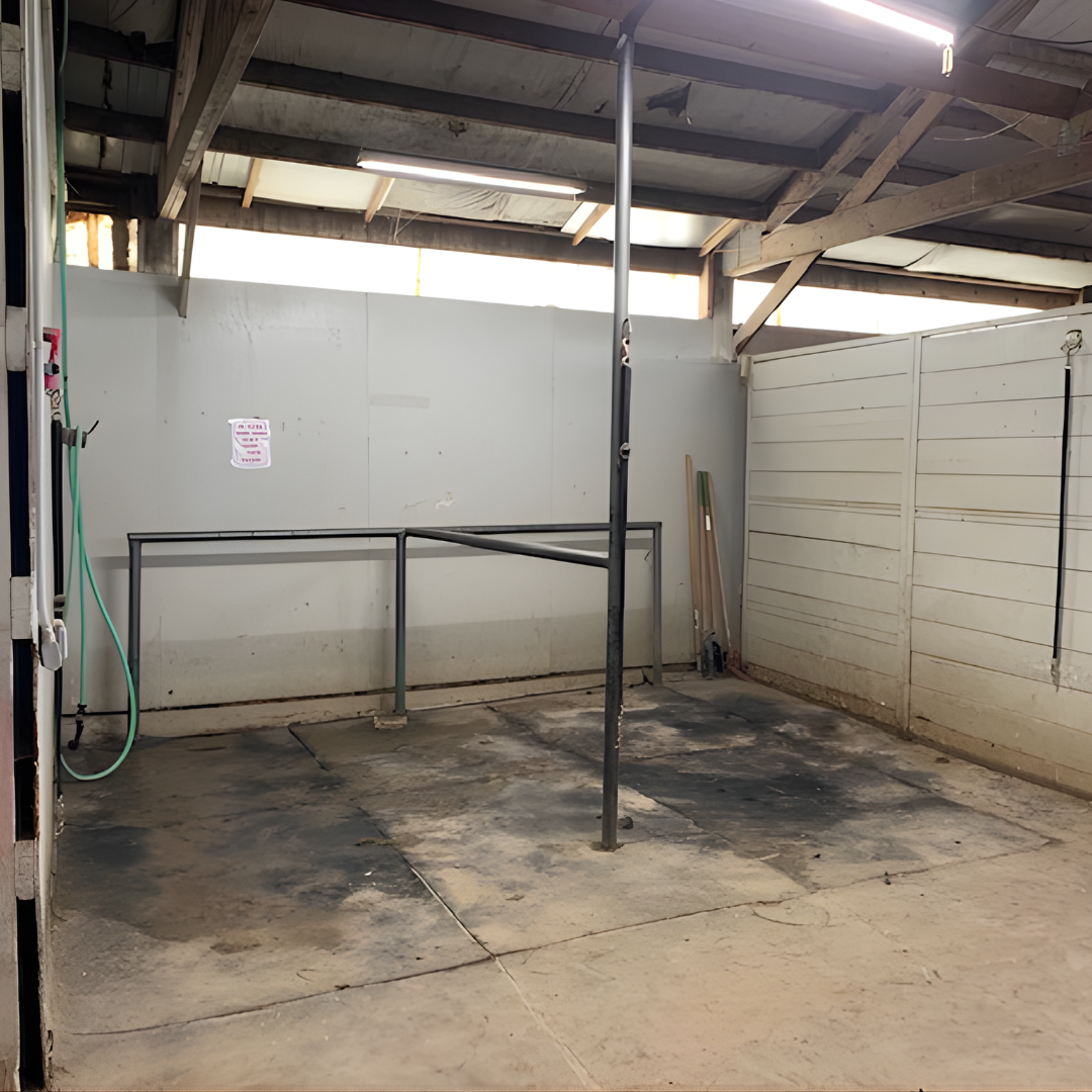 An empty car wash bay with soiled concrete floor, a water hose on the left side, and white wooden privacy fences on the sides, with some building materials leaning against the back wall.