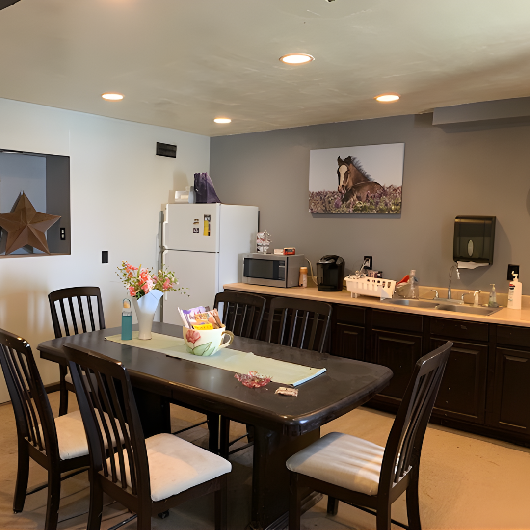 A dining area with a dark wooden table surrounded by six matching chairs with cushioned seats. On the table, there is a white vase with pink flowers, a decorative bowl, a small bottle, and some paper clips. The background features a kitchenette with a microwave, coffee maker, refrigerator, and a sink. A picture of a horse hangs on the wall, and there is a paper towel dispenser mounted near the sink.