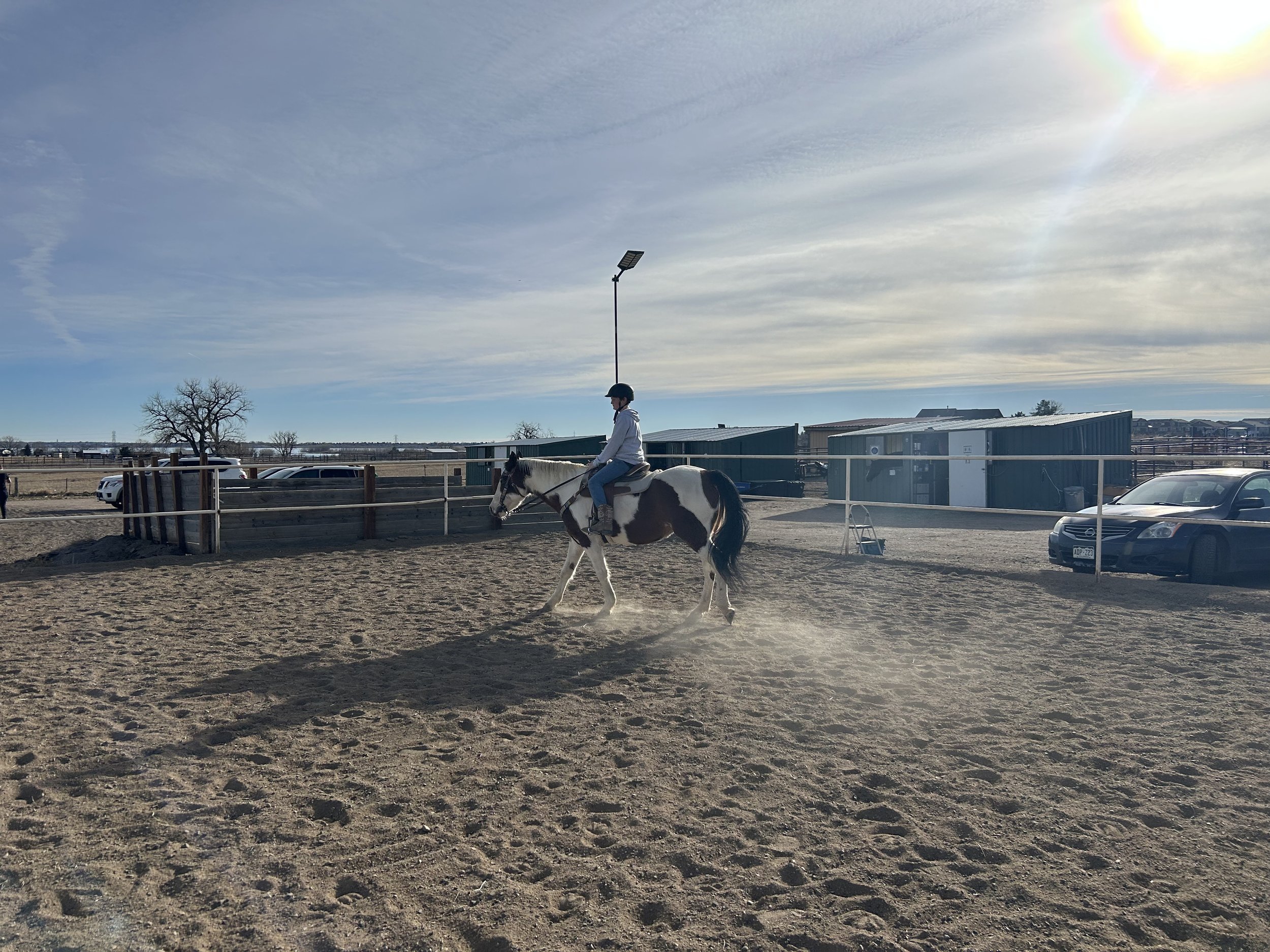 Person riding a pinto horse in an outdoor arena with a fence, parked cars, and storage buildings in the background on a sunny day.