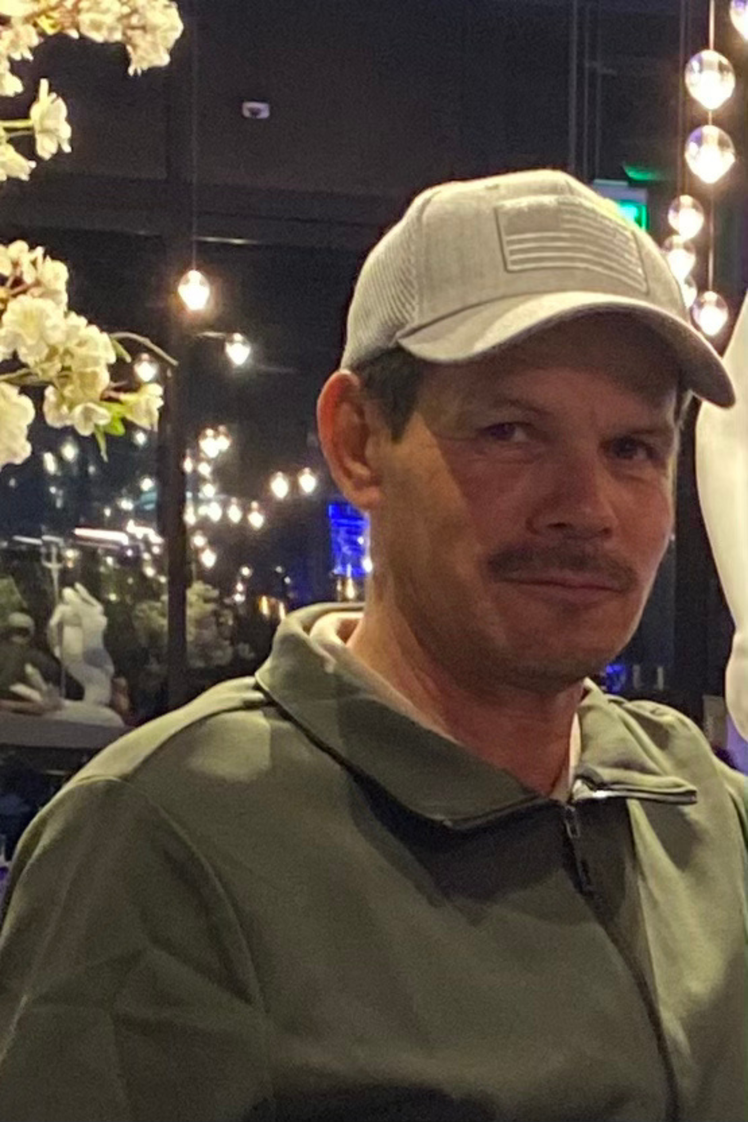 A man wearing a light gray baseball cap and gray jacket sitting indoors near a decorative display with flowers, with string lights and a dark background visible behind him.