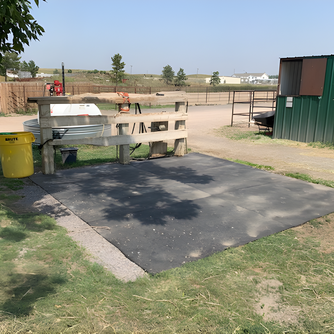 A small outdoor area with a black rubber mat on the ground, surrounded by grass, with a wooden railing, a cold plunge tub, a yellow trash bin, and some trees in the background in a rural setting.