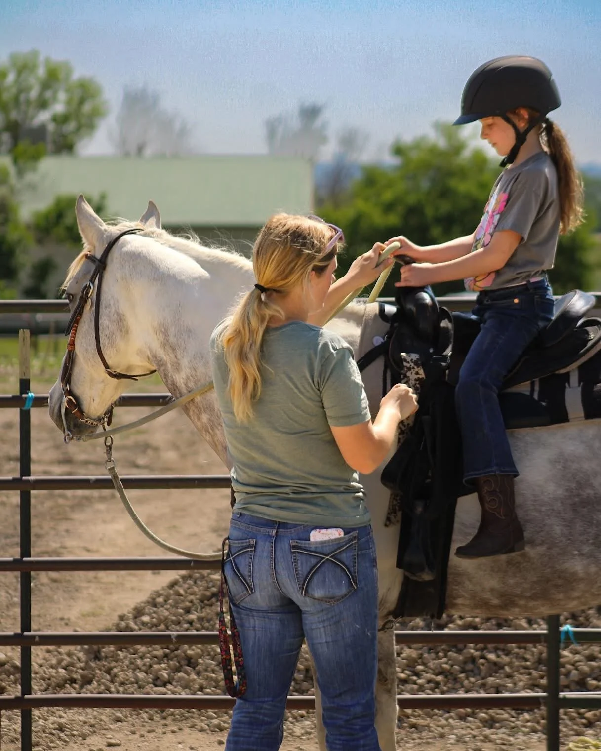 A young girl sitting on a white horse with a black saddle and helmet, having her riding gear checked by a woman standing beside the horse. The scene takes place in an outdoor riding arena with green trees and a building in the background.