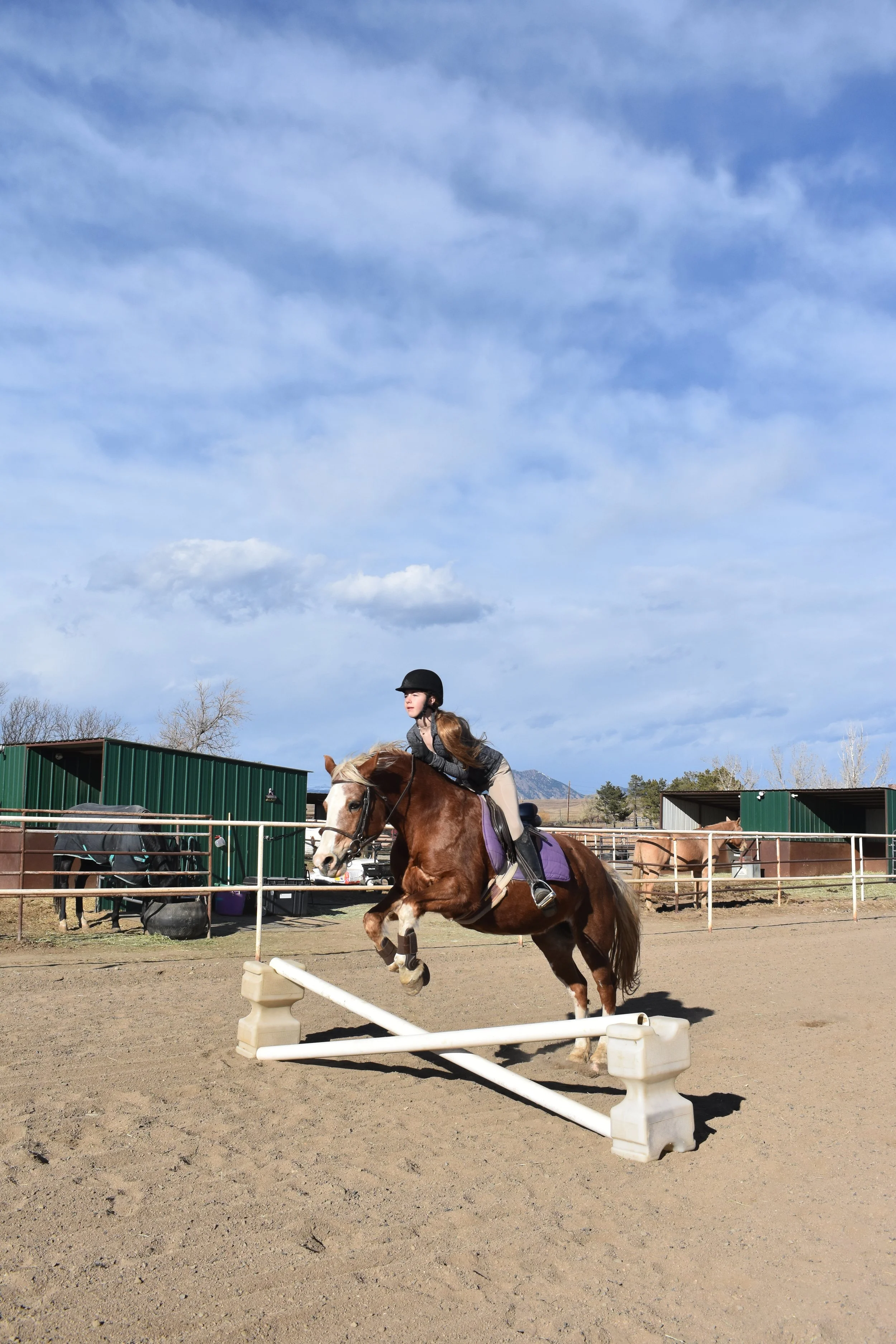Young girl riding a brown and white horse as it jumps over a small obstacle in an outdoor riding arena with green barns and blue sky in the background.