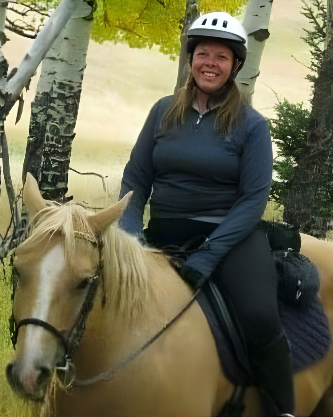 A woman smiling while riding a light-colored horse in a wooded area with trees and green foliage in the background, wearing a helmet and outdoor clothing.