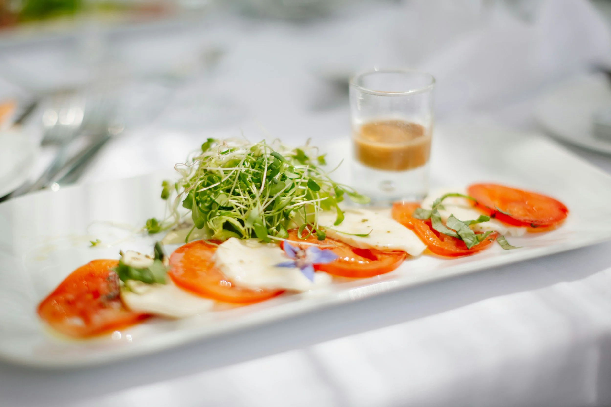 Plate with sliced tomatoes, mozzarella cheese, and leafy greens, accompanied by a glass of dressing or sauce on a white tablecloth.