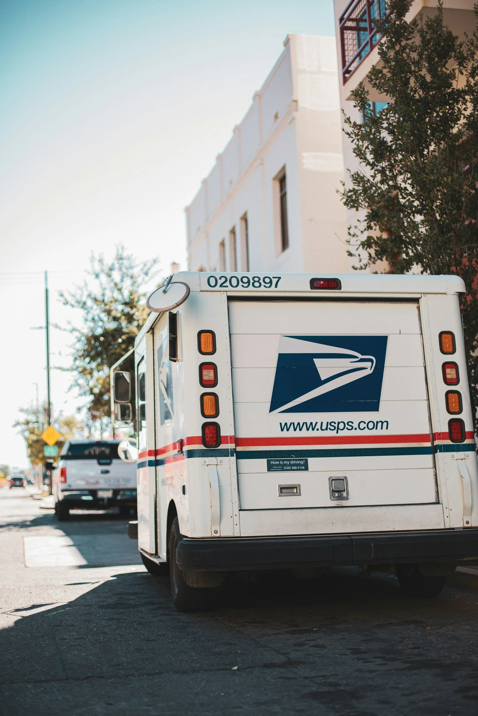 The back of a USPS delivery truck on a city street with apartment buildings and trees in the background.