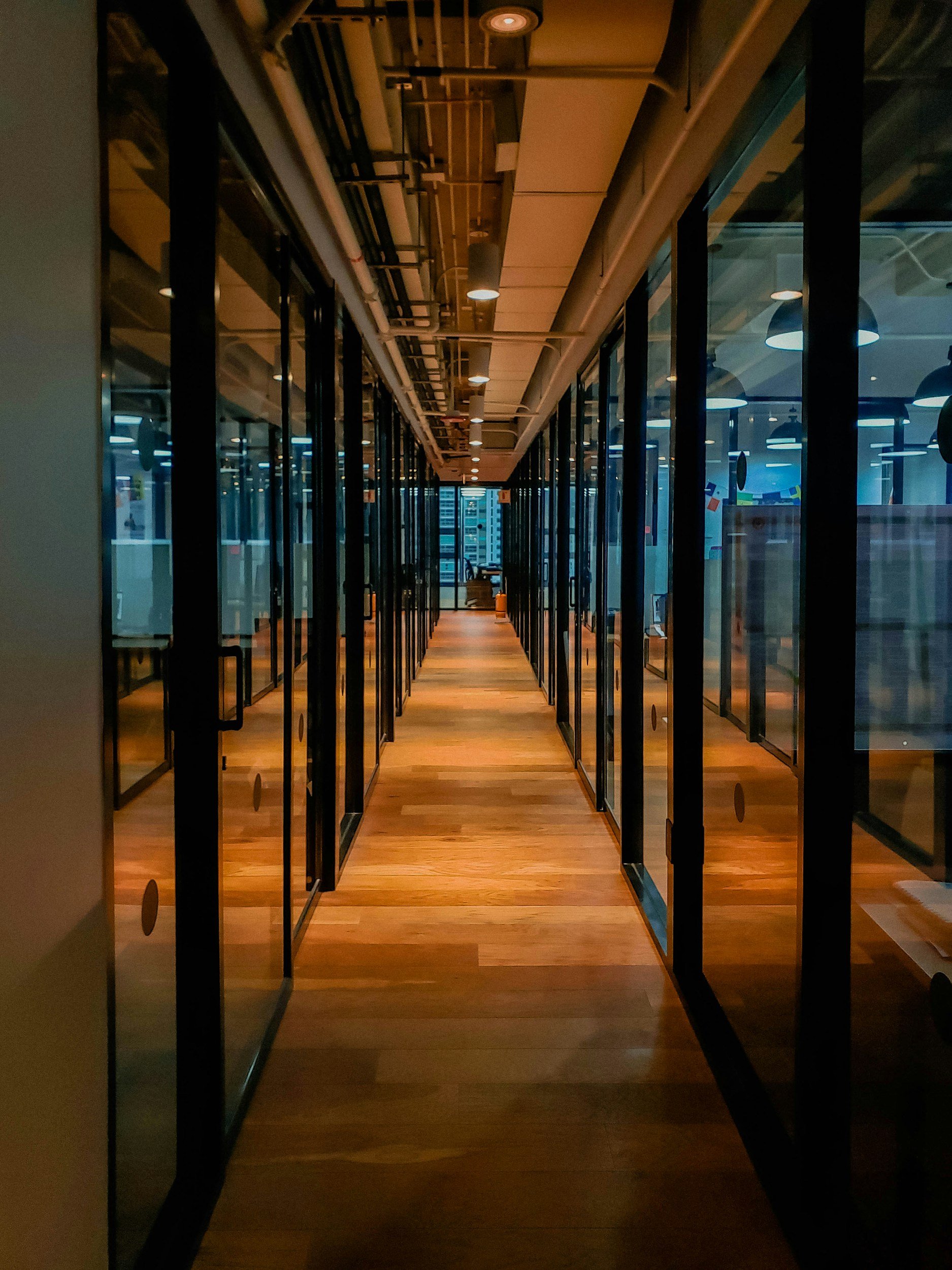 A hallway with glass walls on either side, revealing an office space with desks and chairs. The floor is wooden, and the ceiling has visible piping and lighting.