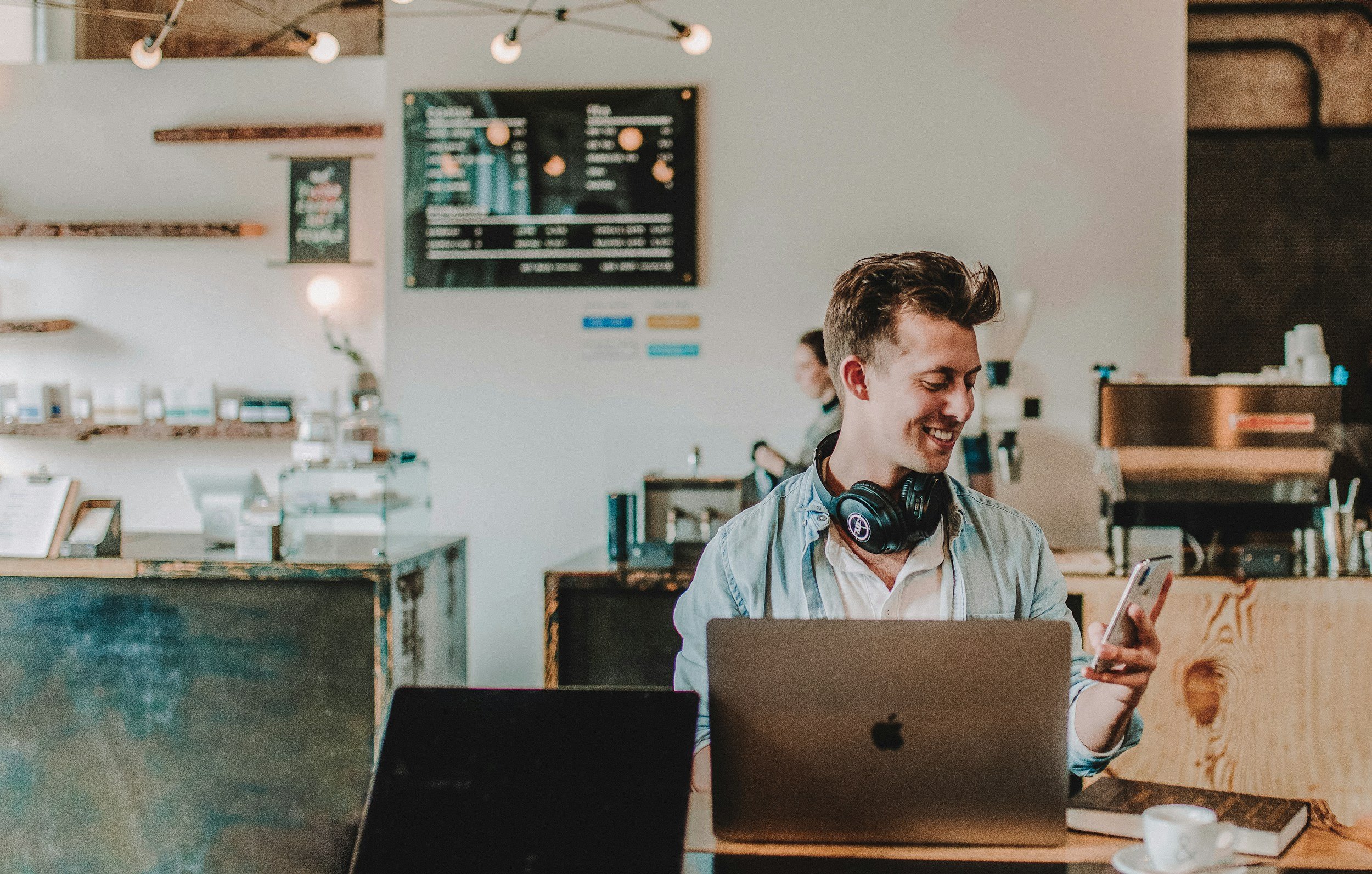 A smiling young man with headphones around his neck looking at his phone, working on a laptop in a cafe.