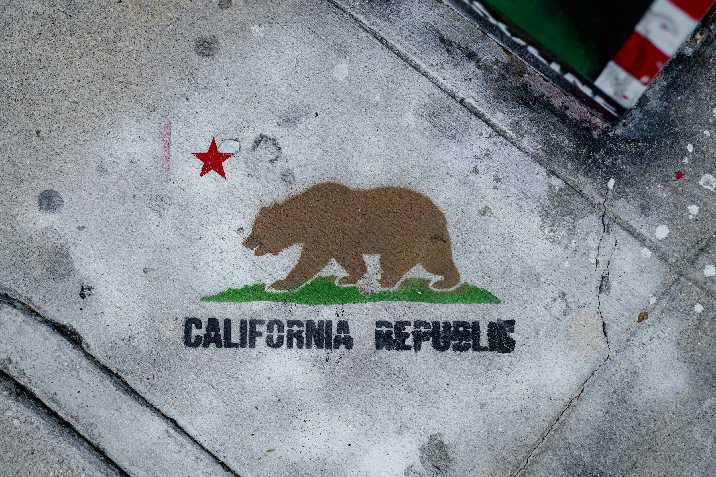 Street art of a bear silhouette outlined in brown, with a green base, painted on the sidewalk. Red star above the bear and black text below reading 'California Bear'.