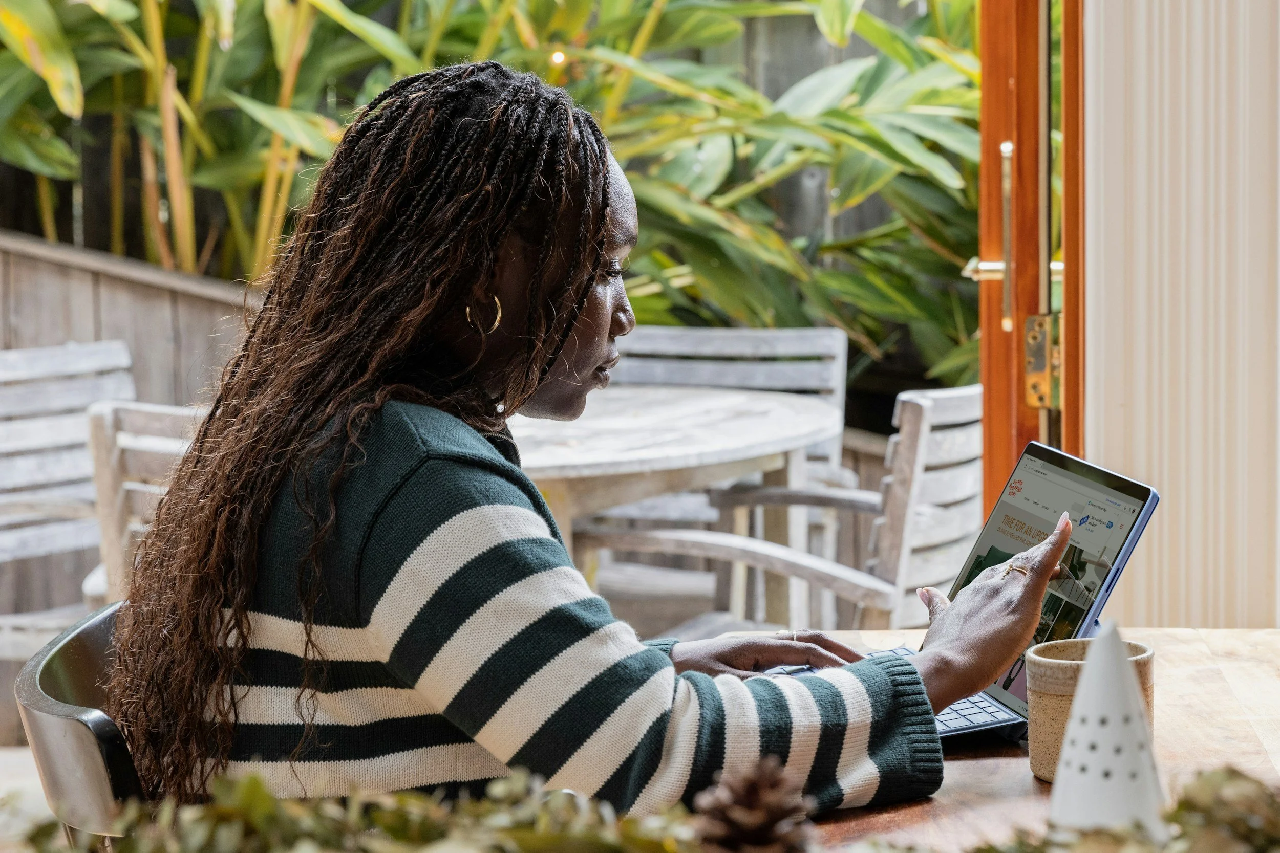 Woman with long, curly hair sitting at a table using a tablet, with a background of leafy plants and a wooden fence.