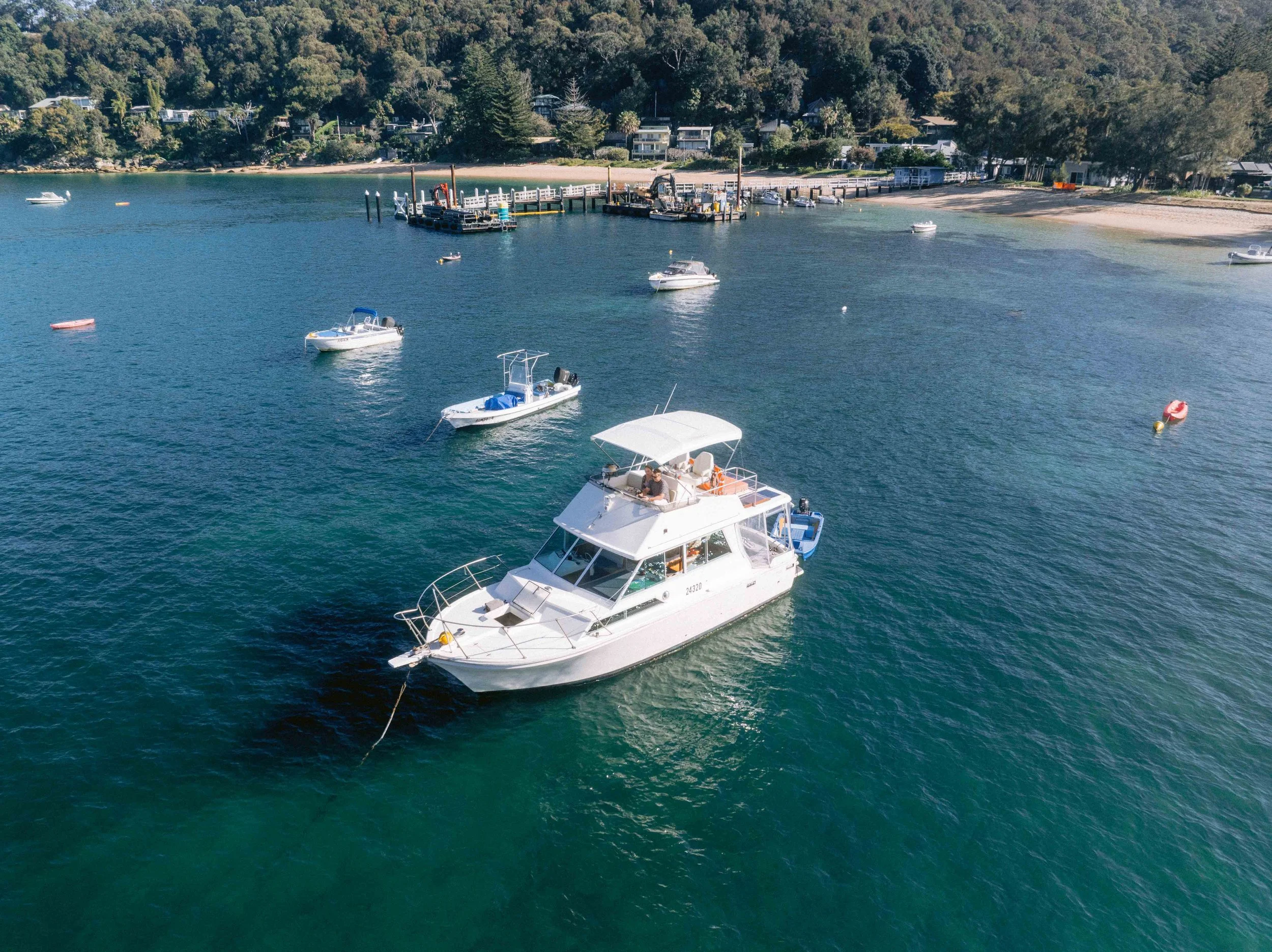 Aerial view of boats docked in a marina near a wooded shoreline with houses, sandy beach, and calm water. Church Point Charter self-drive charter boat hire Pittwater
