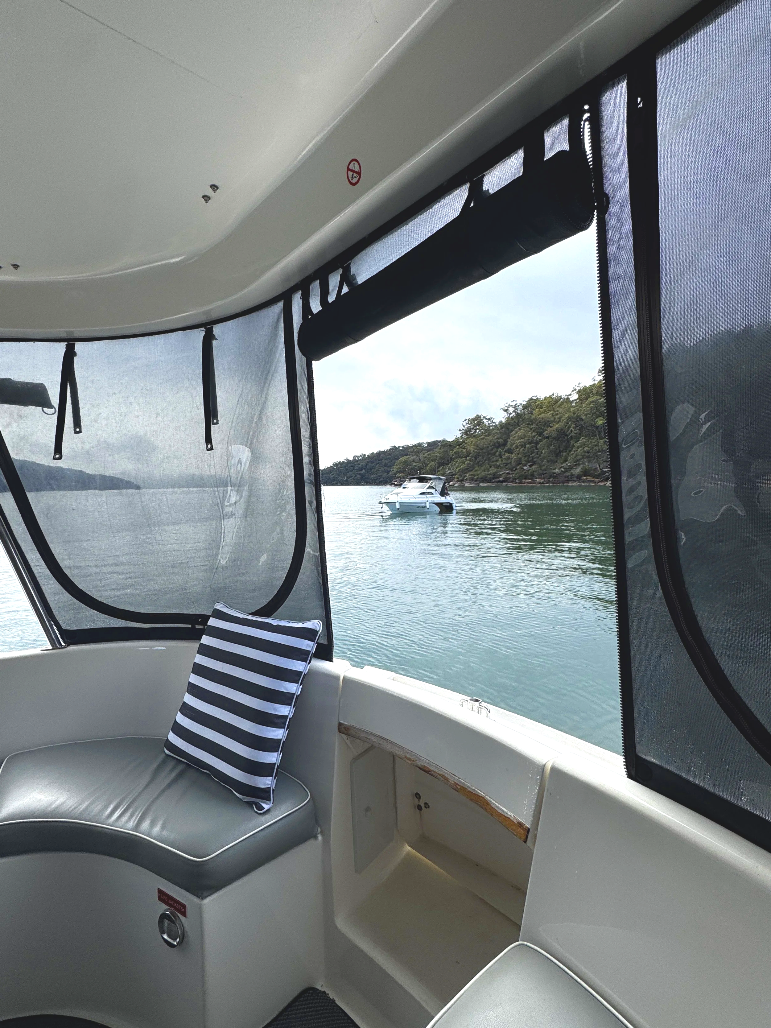 View from inside a boat looking out through screened windows at water and a distant boat. The interior has a curved seating area with a striped pillow, and there are zippers and a small storage compartment inside. Church Point Charter self-drive char