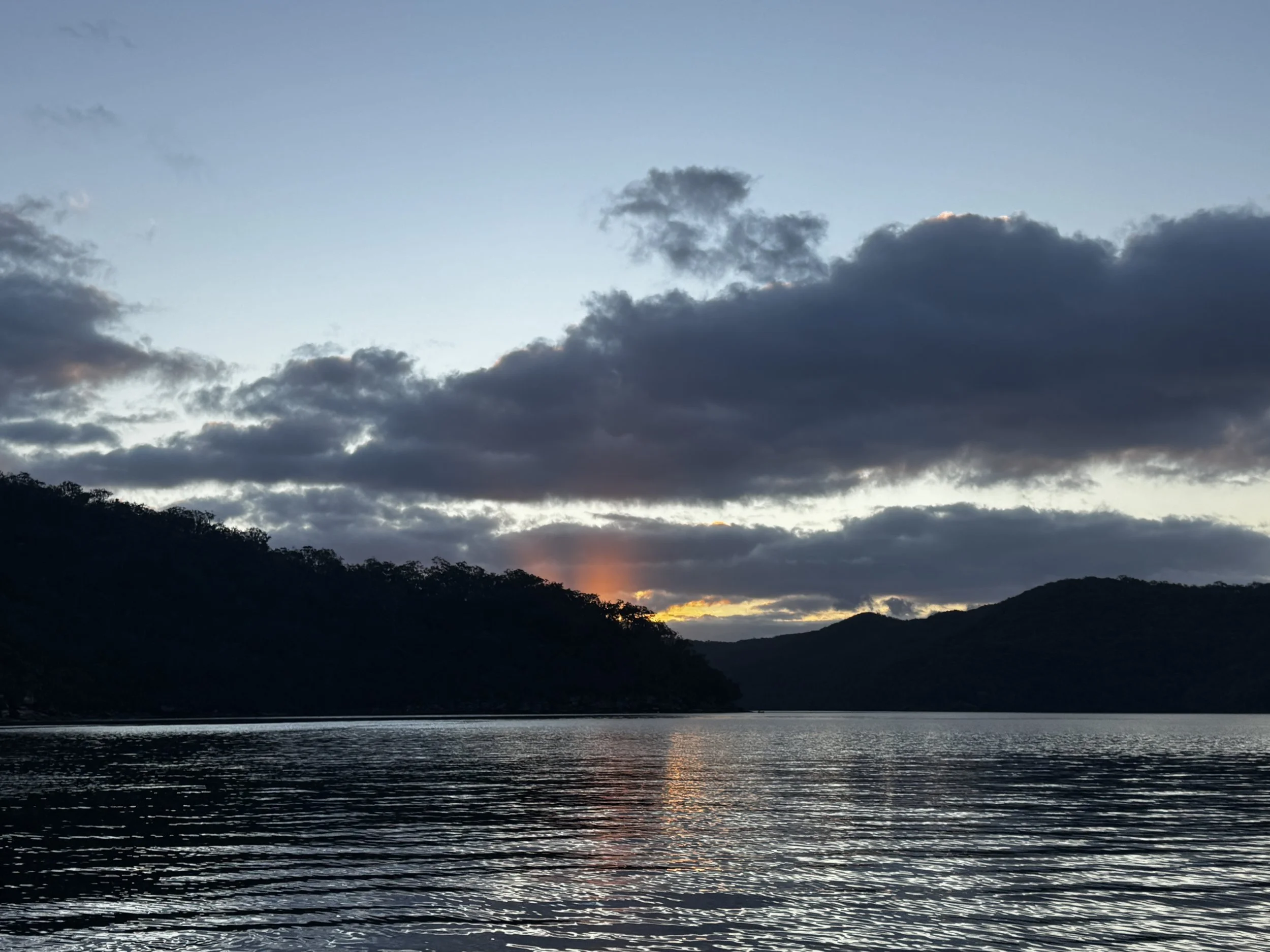 View of a river or lake with reflections of surrounding hills and a sunset or sunrise sky with clouds. Church Point Charter self-drive charter boat hire Pittwater