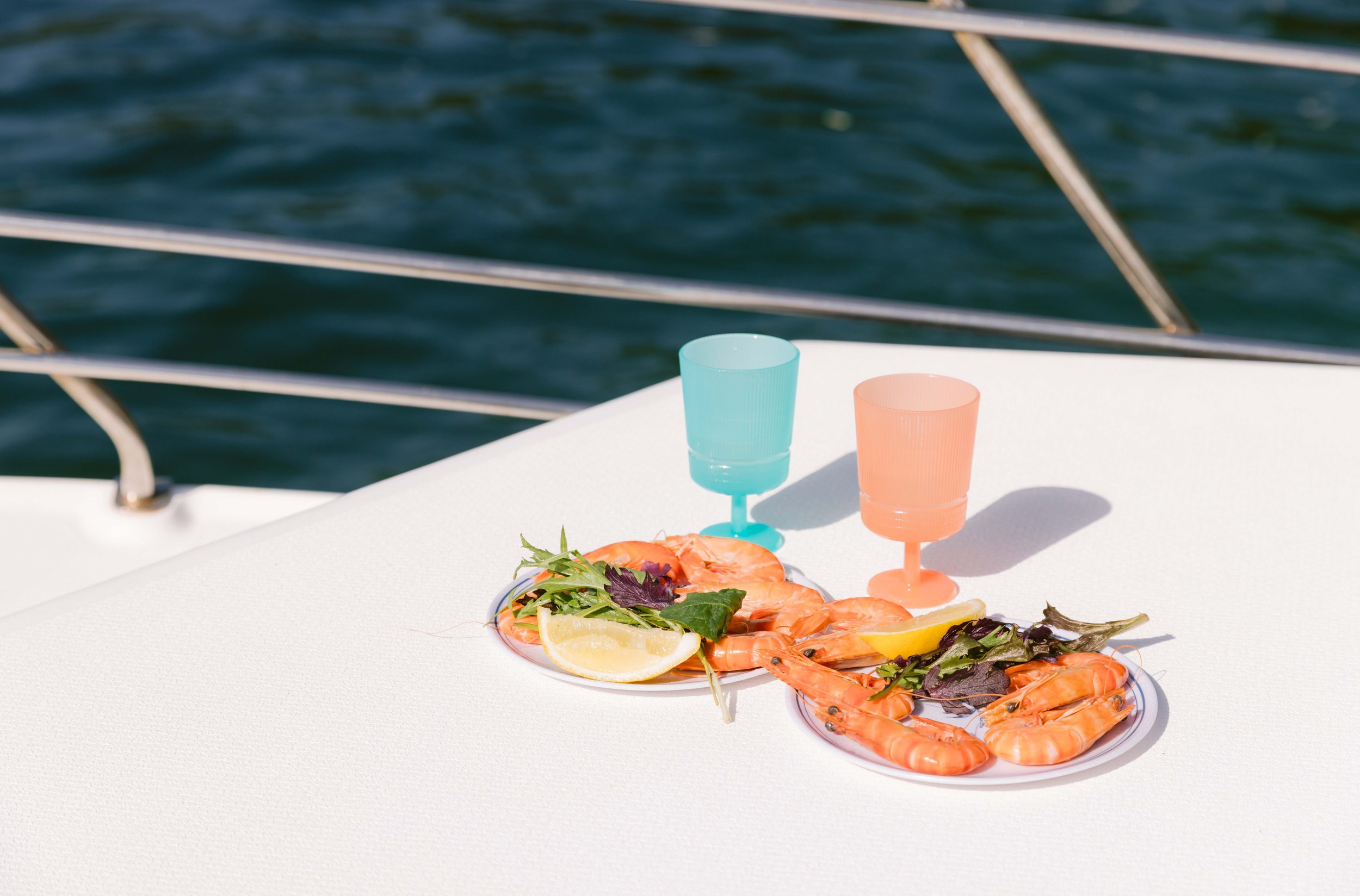 A plate of smoked salmon with lemon slices, mixed greens, and herbs, with two colorful plastic cups, set on a white surface near a body of water with boat railing in the background. Church Point Charter self-drive charter boat hire Pittwater