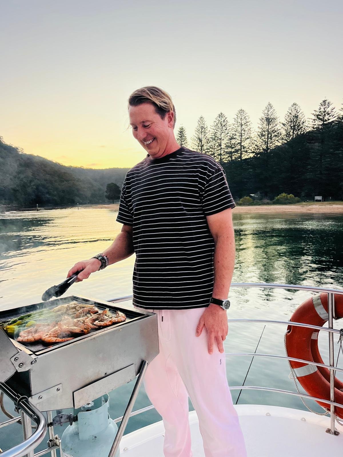 A smiling man in a black and white striped shirt grilling seafood on a boat at sunset, with a body of water and trees in the background. Church Point Charter self-drive charter boat hire Pittwater