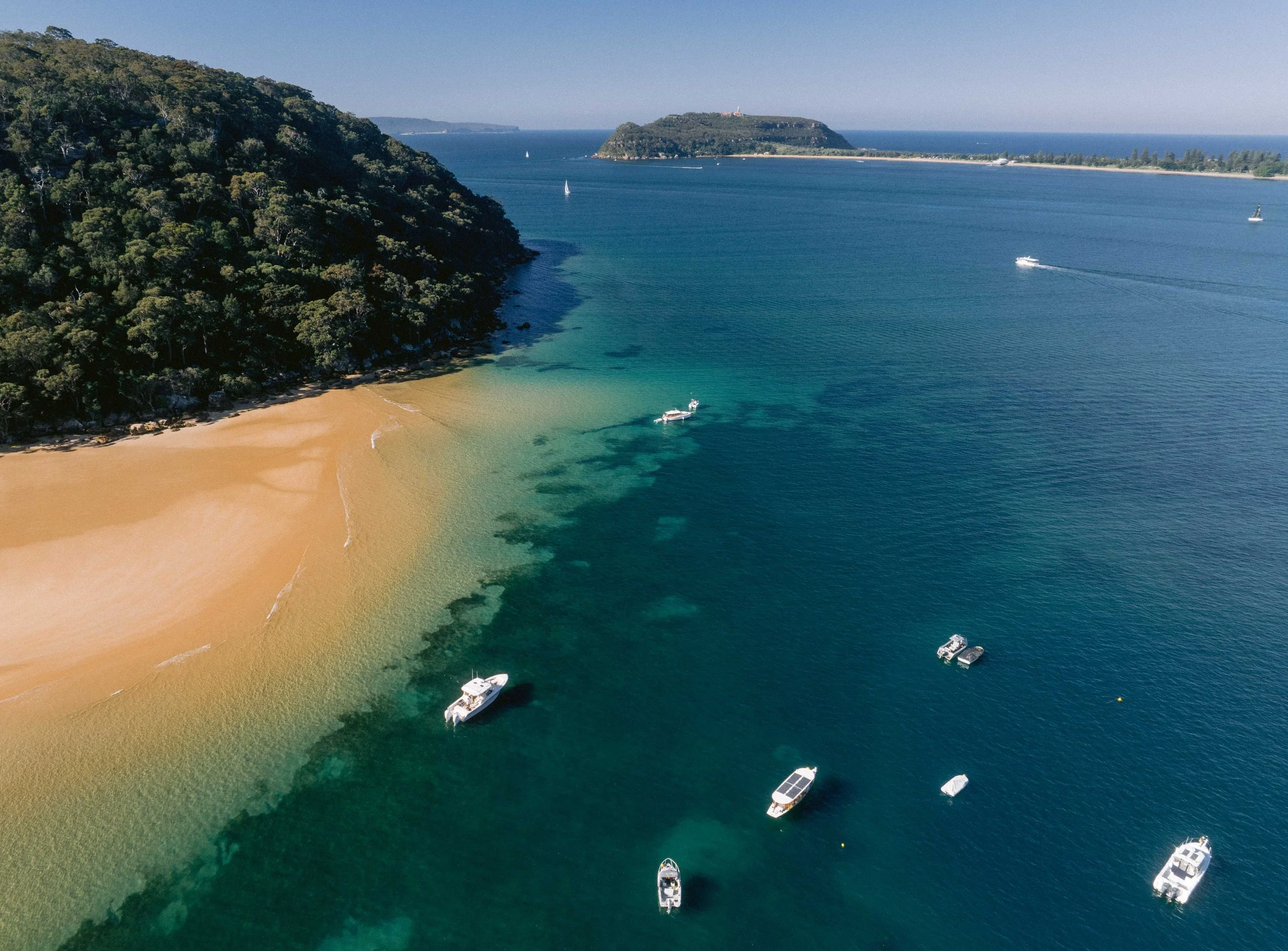An aerial view of a coastline with a sandy beach, clear turquoise water, and a forested hill on the left. Several boats are anchored and sailing in the water, with offshore islands in the background.