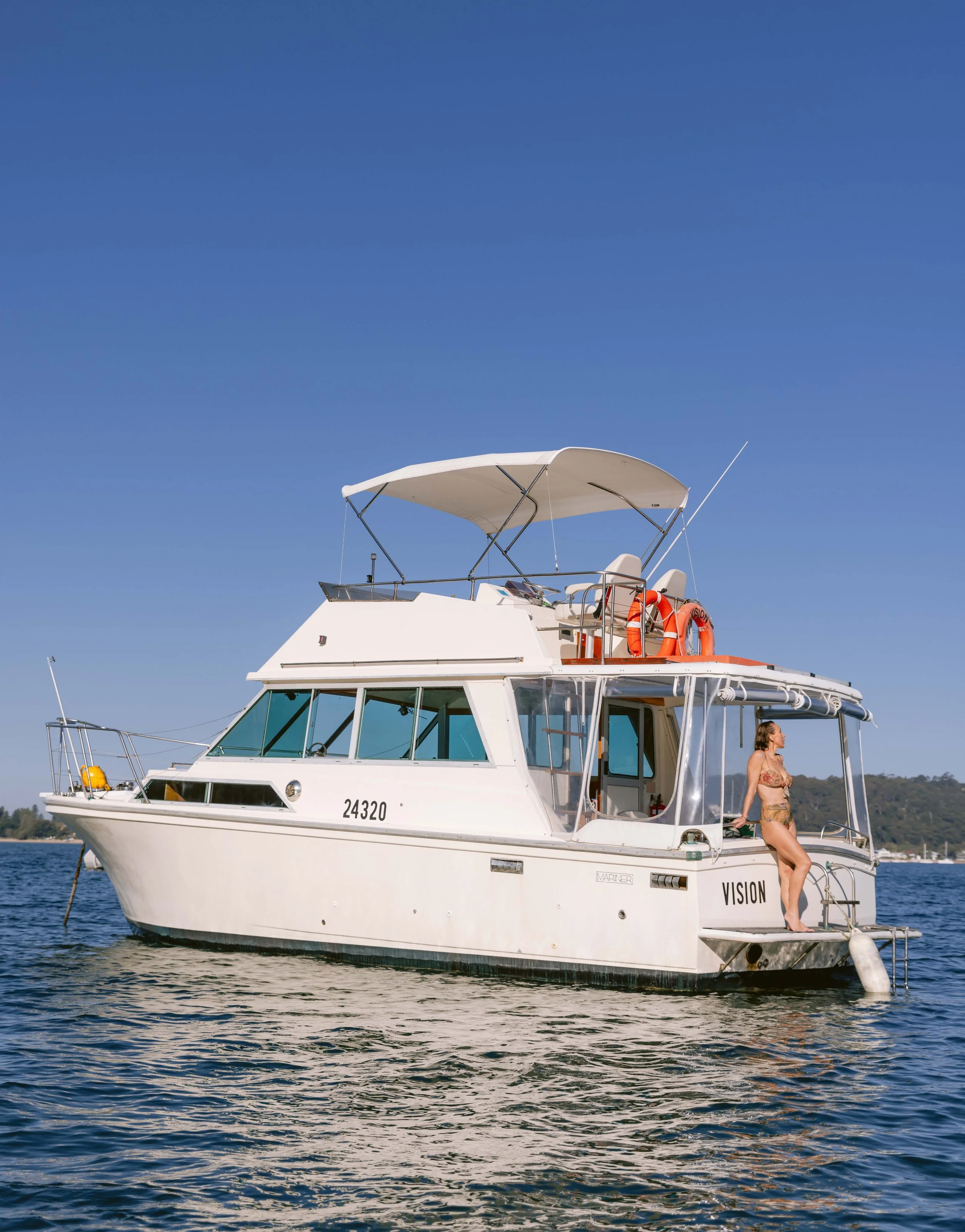 A woman in a swimsuit standing on the back of a white yacht named 'VISION' on a body of water, with a blue sky and distant shoreline in the background. Church Point Charter self-drive charter boat hire Pittwater