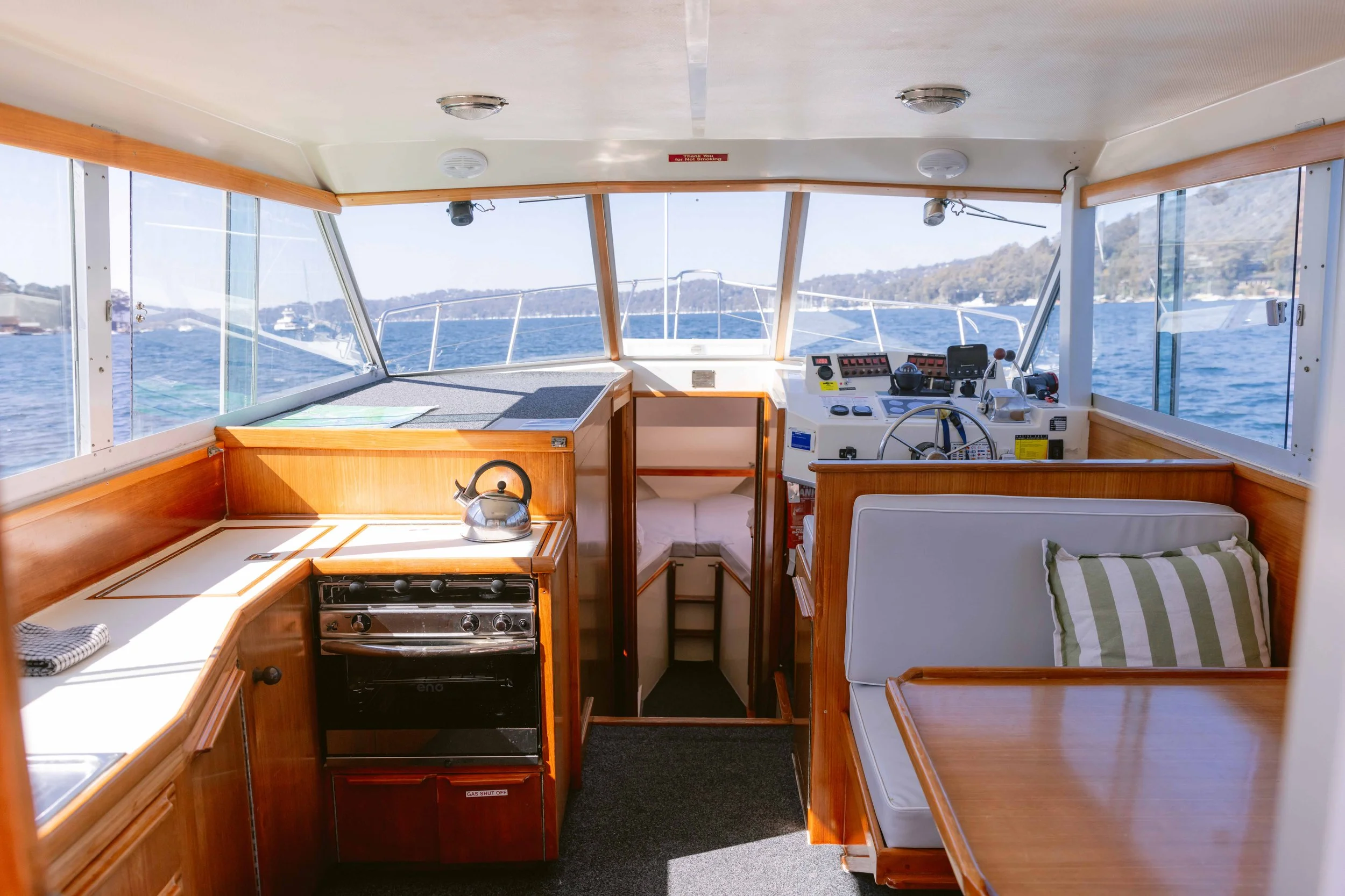 The interior of a boat cabin with wood furnishings, a small stove, a bench with a striped pillow. Church Point Charter self-drive charter boat hire Pittwater