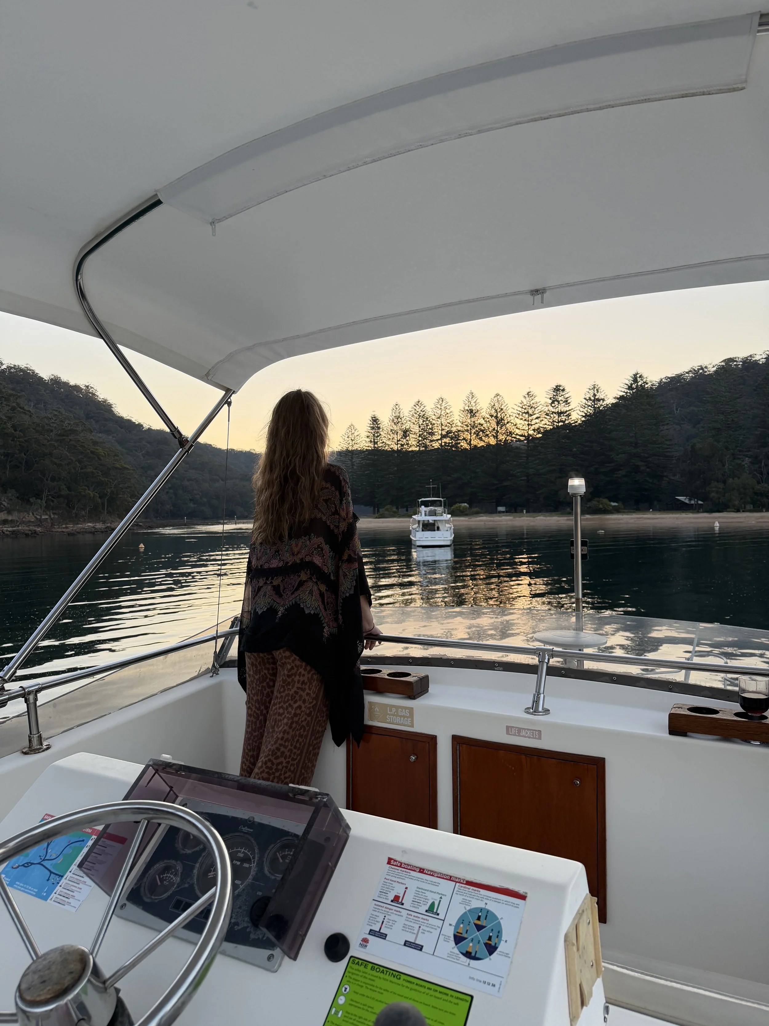 A woman with long hair stands on a boat, looking at the water and a boat in the distance during sunset. The boat has a white canopy and various safety and navigation signs are visible. Church Point Charter self-drive charter boat hire Pittwater