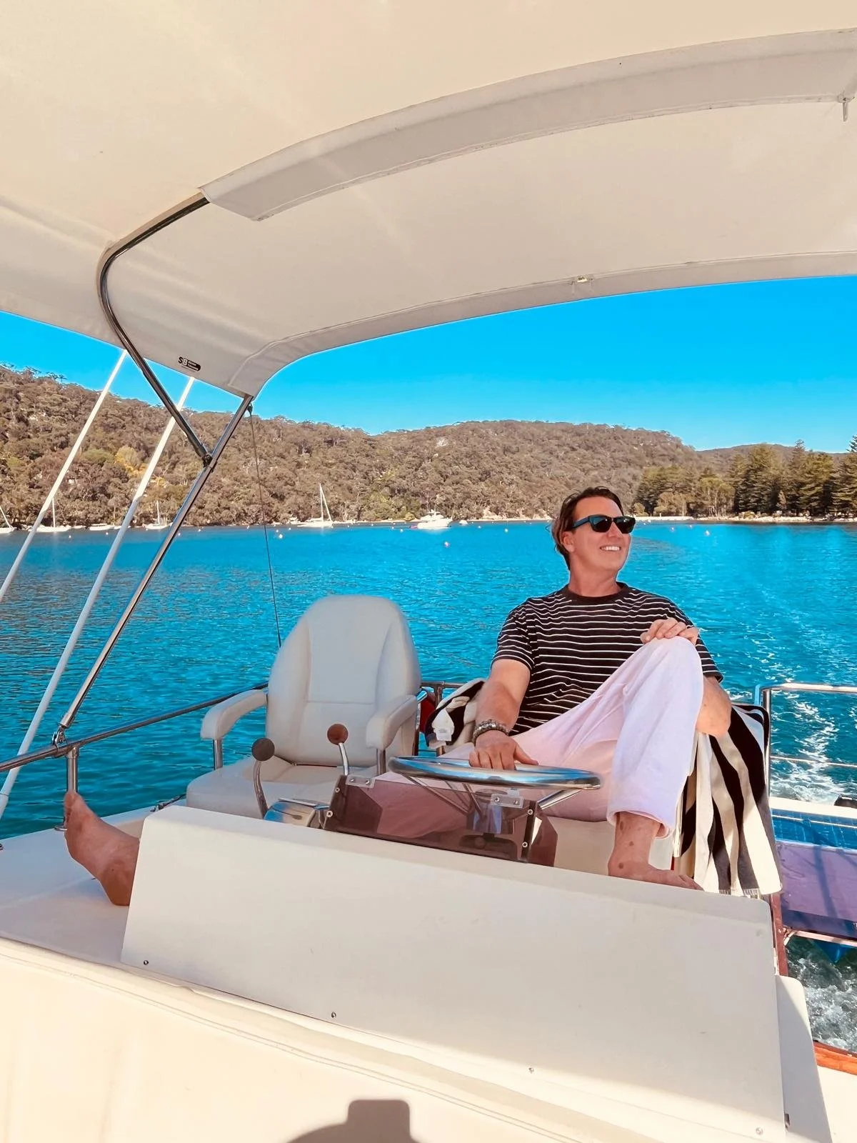 A man sitting on a boat enjoying a sunny day on a body of water with hills and trees in the background. Church Point Charter self-drive charter boat hire Pittwater
