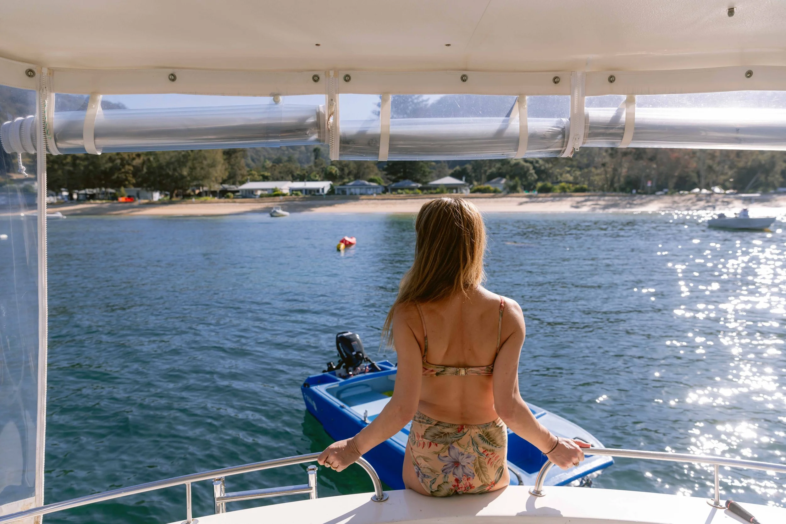 A woman with long blonde hair standing on a boat, looking out at the water with a shoreline, houses, trees, and boats in the background. The boat has a blue railing and a clear plastic cover. Church Point Charter self-drive charter boat hire Pittwate