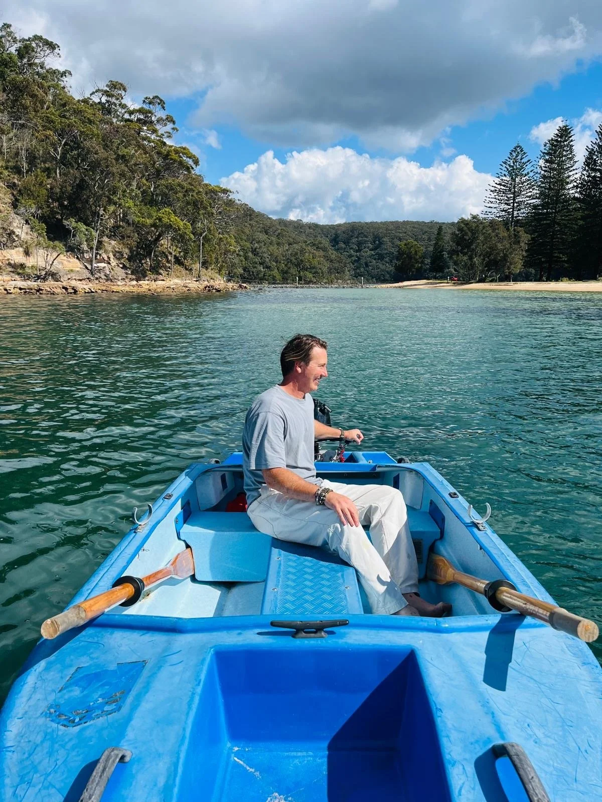 A man in a gray t-shirt and white pants sits in a bright blue rowboat on calm green water, smiling as he looks to his left. The landscape features forested hills, a shoreline, and partly cloudy sky. Church Point Charter self-drive charter boat hire P