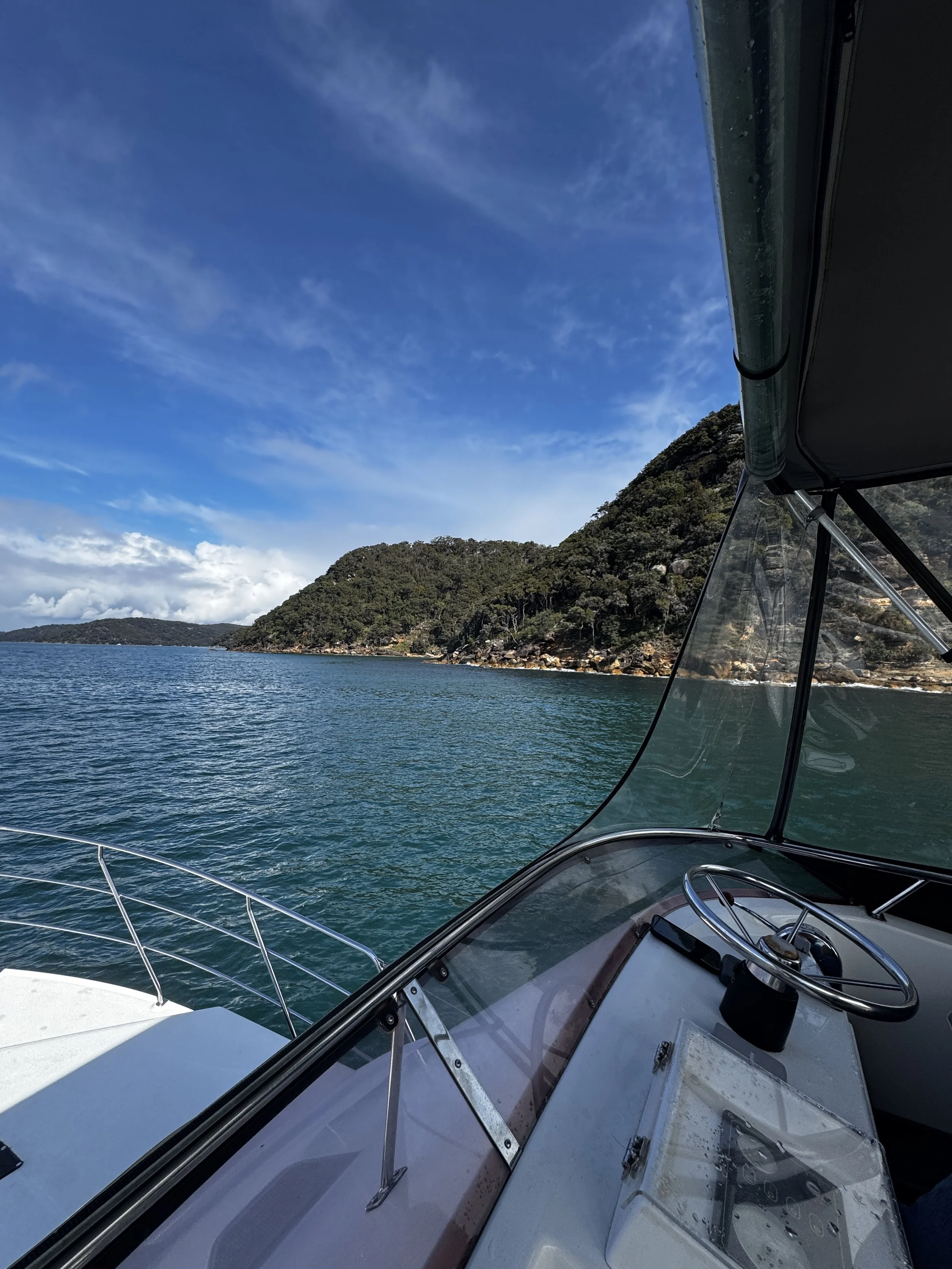 View from a boat showing calm water, wooded hills, and a blue sky with scattered clouds, with part of the boat's cockpit, steering wheel, and windshield visible in the foreground. Church Point Charter self-drive charter boat hire Pittwater