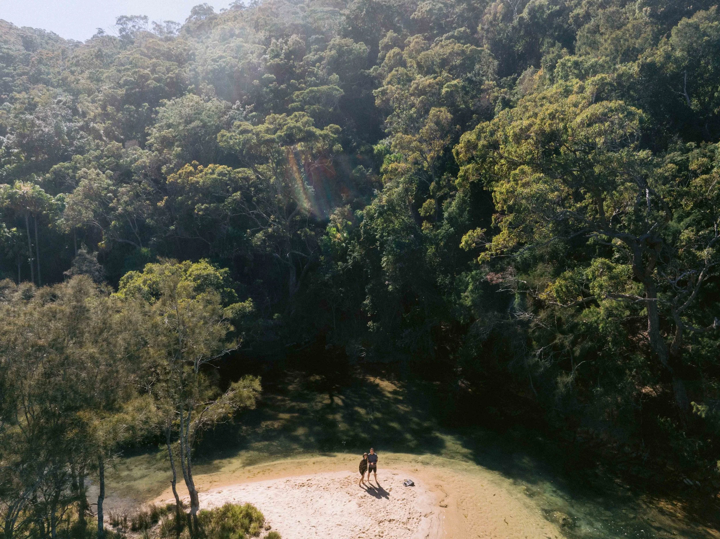 A forested area with two people standing on a small sandy clearing near a body of water, with dense green trees and a mountain in the background. Church Point Charter self-drive charter boat hire Pittwater