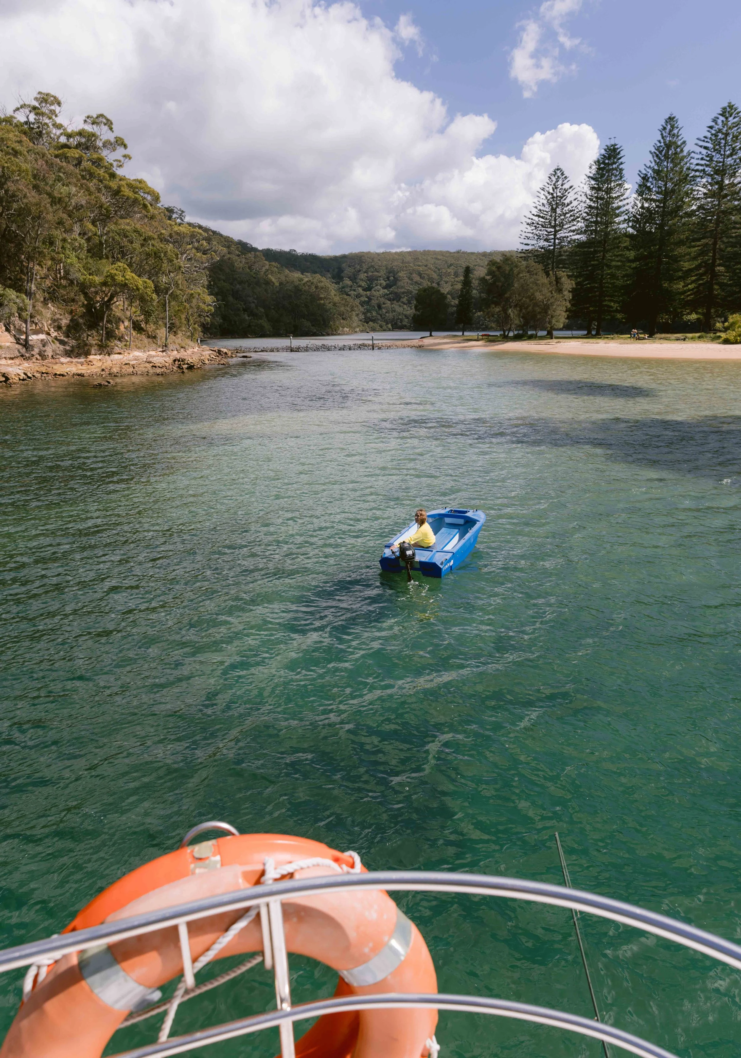 View from a boat on a river with a person sitting in a small blue boat in the water, surrounded by green trees and hills, with a cloudy sky overhead. Church Point Charter self-drive charter boat hire Pittwater