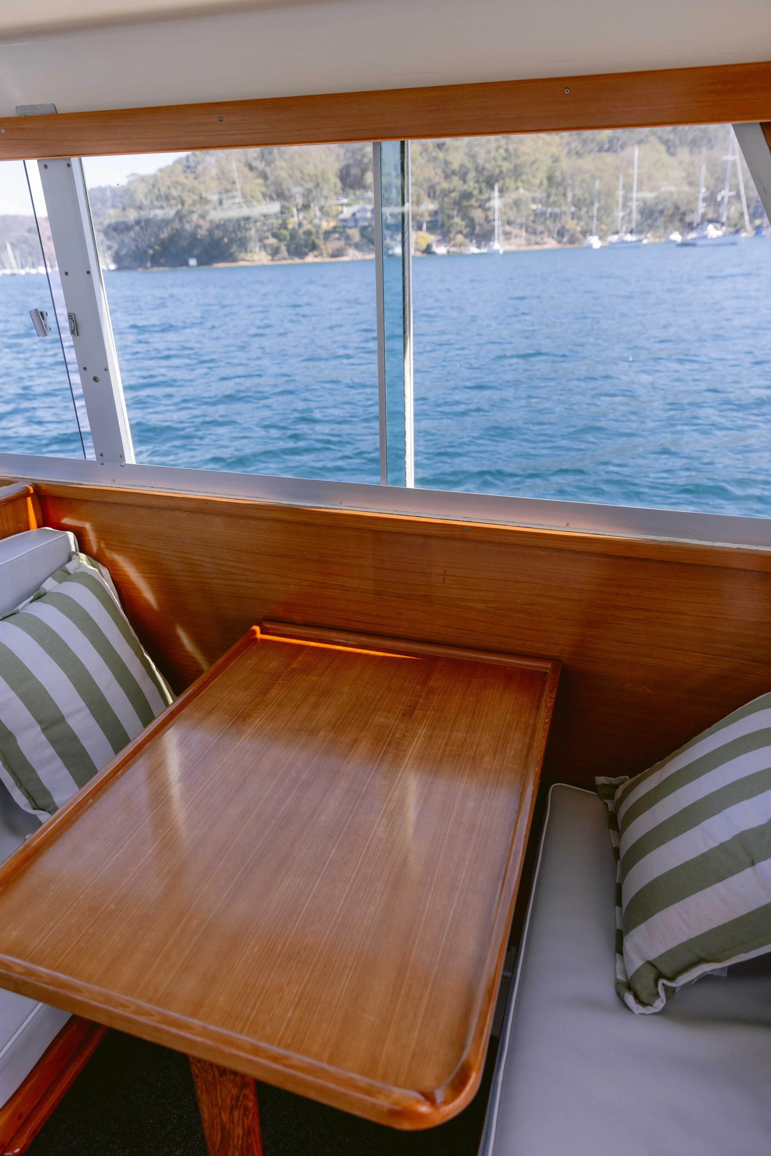 View from inside a boat showing a wooden table with striped pillows against the wall, overlooking calm water and a distant shoreline with trees and anchored sailboats.