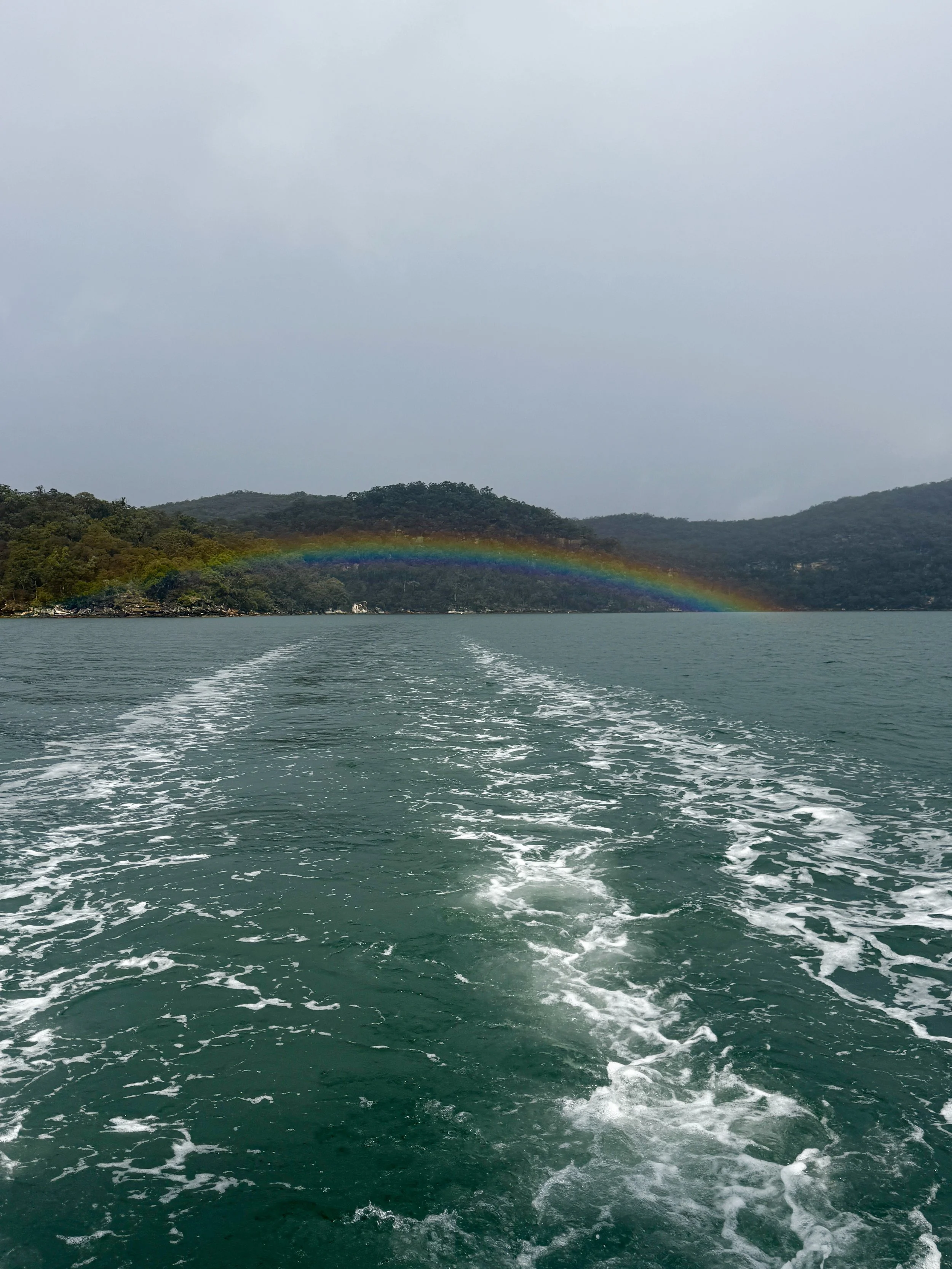 View of a body of water with a trail of boat wake, green hills in the background, and a rainbow spanning across the sky above the hills. Church Point Charter self-drive charter boat hire Pittwater