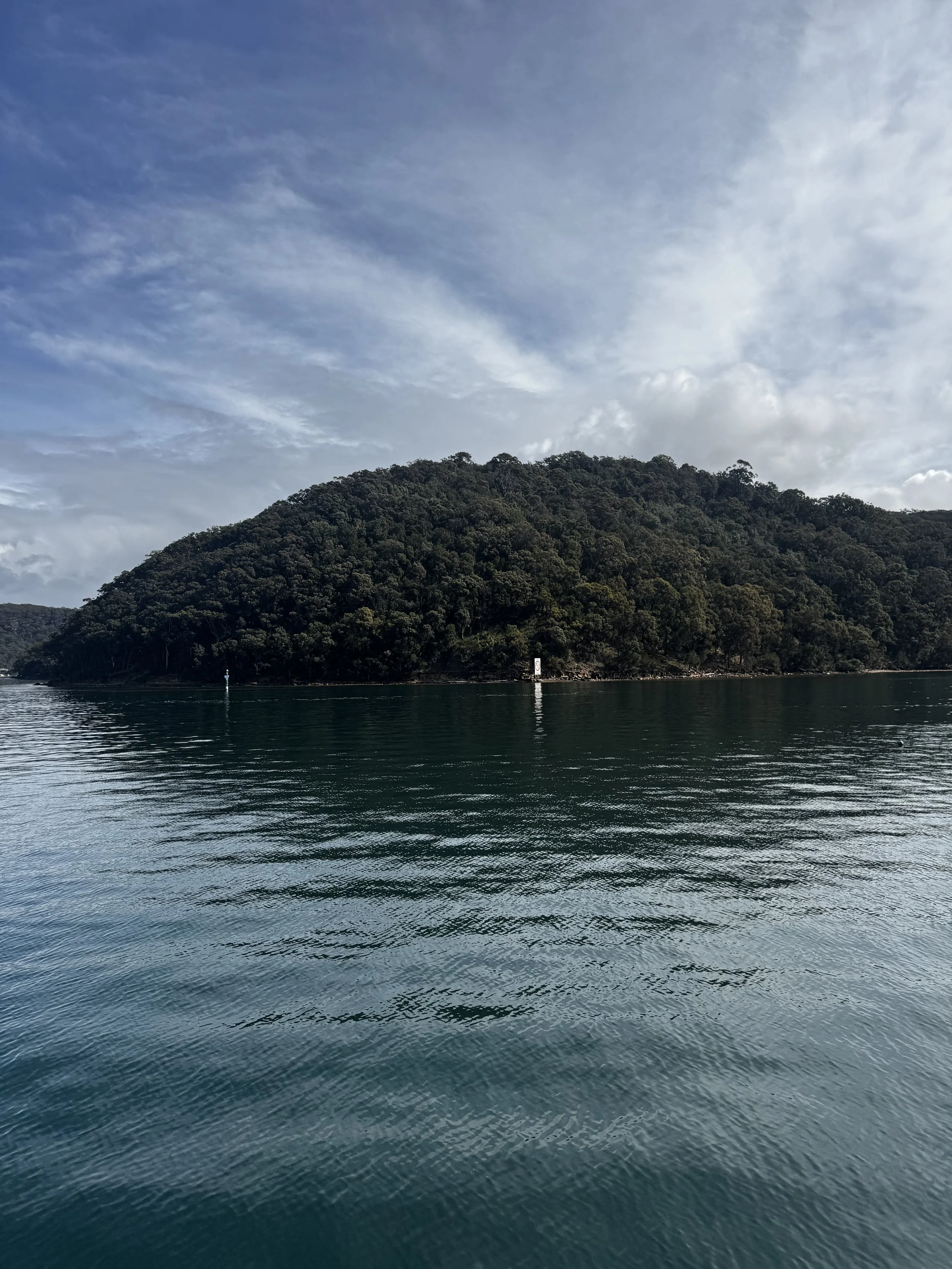 Calm ocean water with reflections and a forested hill in the background under a partly cloudy sky. Church Point Charter self-drive charter boat hire Pittwater