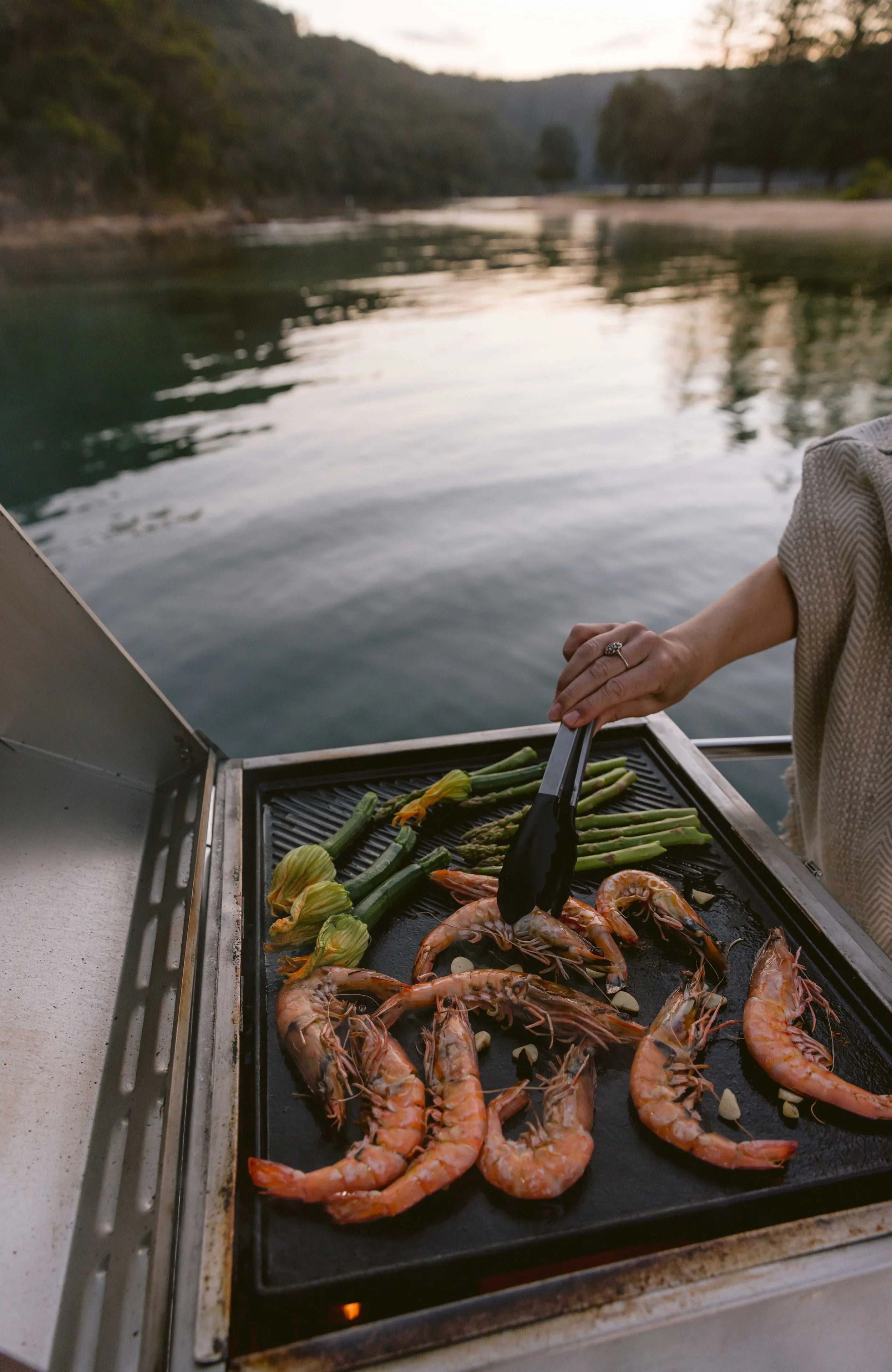 A person grilling shrimp, artichokes, asparagus, and zucchini on a barbecue grill near a river during sunset. Church Point Charter self-drive charter boat hire Pittwater