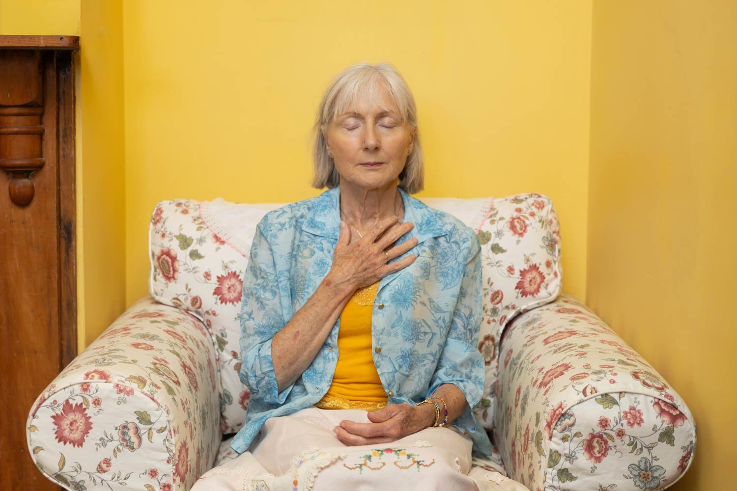 Older lady sitting in an armchair with her hand on her heart, practicing mindfulness