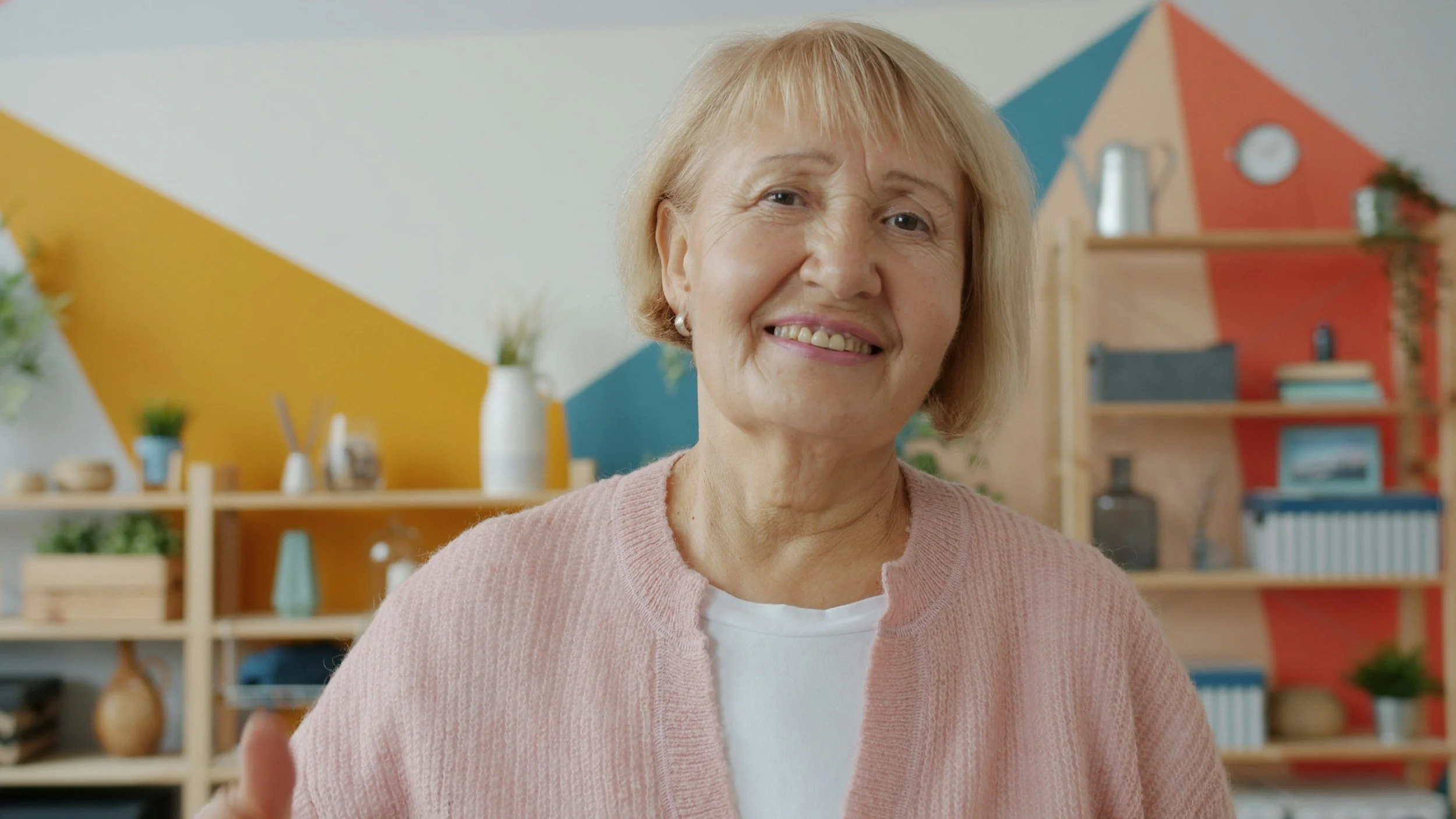Older woman in her studio with a pink cardigan on and a white t-shirt looking hopeful and happy