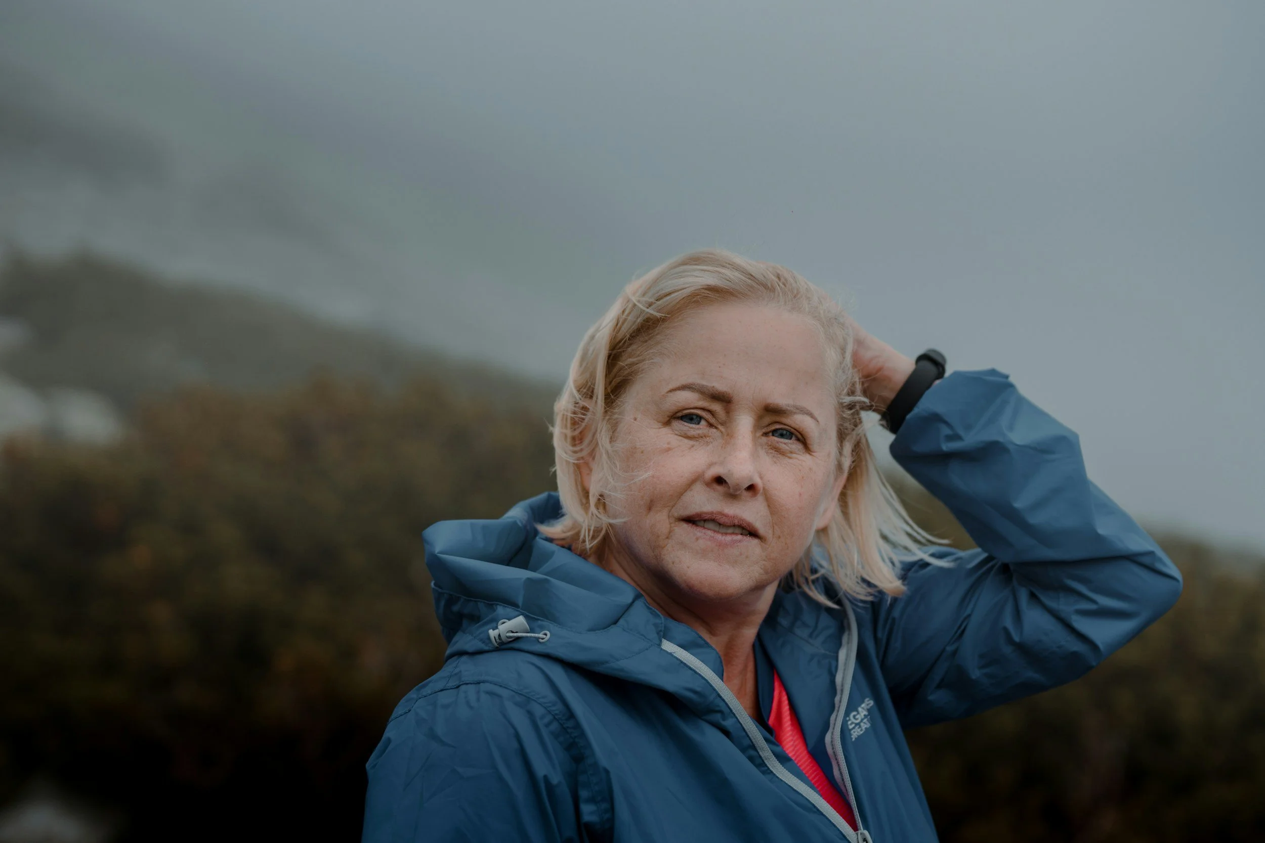 Mature woman  with shoulder length grey hair in blue jacket outside with her arm behind her head looking in the distance. It is a windy day.