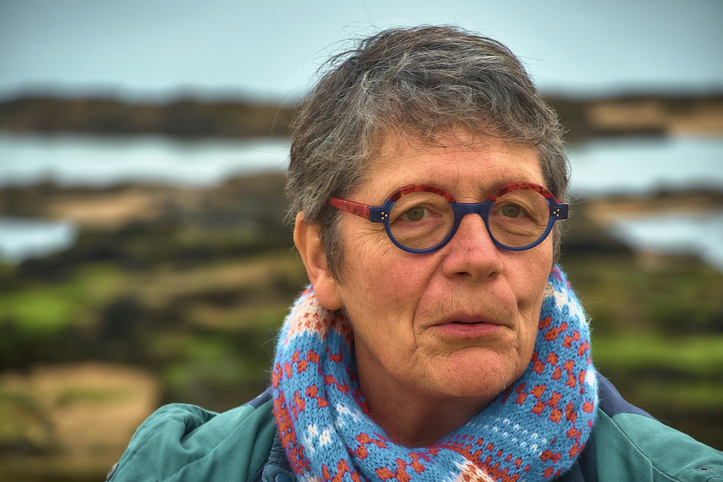 Woman with short grey, browm hair with round glasses and a bright knitted scarf outside near the sea, looking sad.