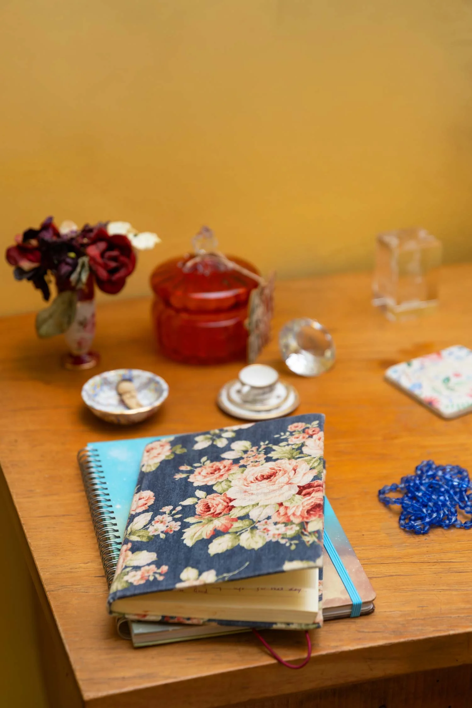 A wooden table with notebooks, a floral fabric pouch and various decorative items including a vase with dried flowers, a red jar, glass ornaments, a jewelry dish, and a cloth wallet.