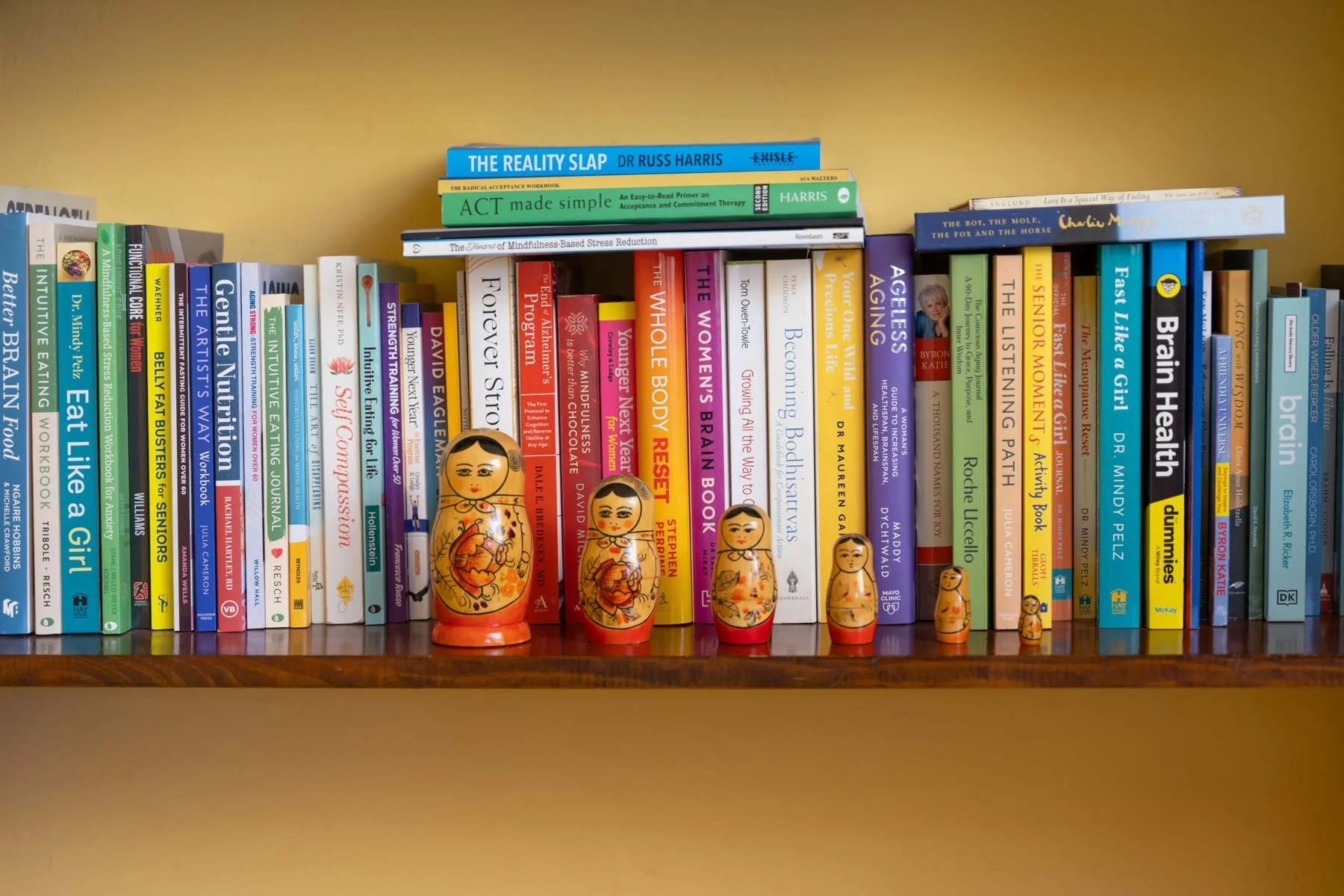 A row of colorful books on a wooden shelf, with five traditional nesting dolls in front of them.